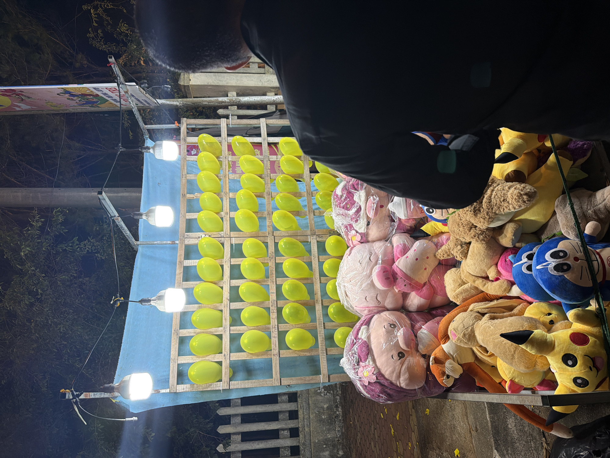 This image depicts a vibrant night scene at an outdoor carnival or fair, likely in Măng Đen, Vietnam. A person, visible from the back and shoulder, wearing a dark jacket, stands in the foreground, seemingly observing or participating in a game.
The central focus is a balloon game stall. Numerous bright yellow balloons are neatly arranged in rows on a wooden grid structure, set against a light blue fabric backdrop. The area is brightly illuminated by several strong white lights mounted on stands, indicating the nighttime setting.
To the right of the game stall, a large collection of plush toys is stacked high, serving as prizes. These include popular characters such as Pikachu and Doraemon, alongside various other animal figures like pigs, rabbits, and teddy bears. Some of these prizes are still encased in clear plastic wrapping.
In the upper left portion of the image, a colorful banner featuring cartoon characters is partially visible. Text on this banner clearly reads "TRÒ CHƠI" (Game), "PHẦN THƯỞNG" (Prizes), and "ĐỒ CHƠI" (Toys), providing further context for the game stall. The dark surrounding environment, contrasted with the brightly lit stall, highlights the evening or night time of the event. The scene conveys an atmosphere of lively entertainment typical of a night market or festival.