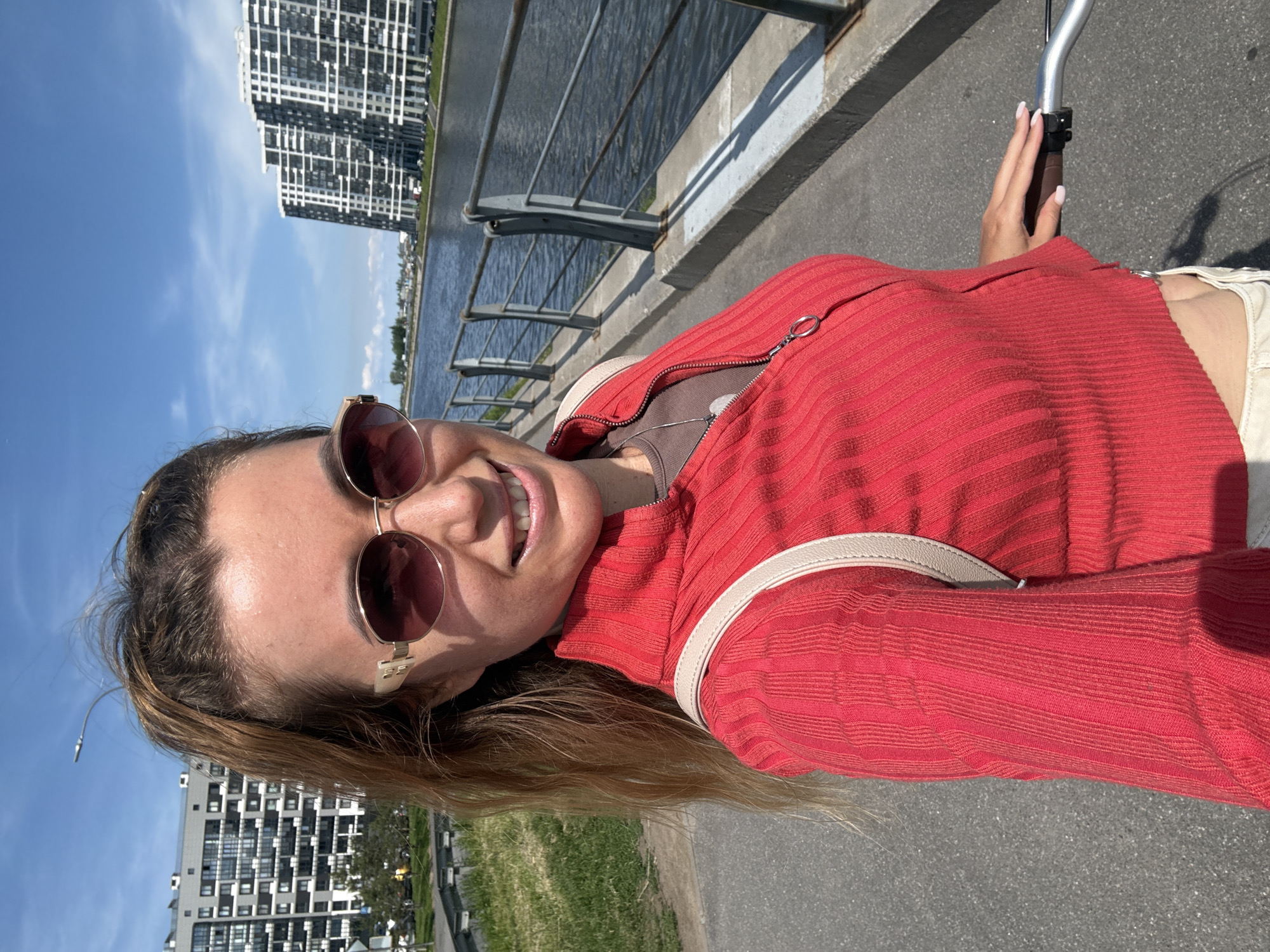 A young woman with blonde hair pulled into a ponytail and wearing oversized sunglasses, a red ribbed zip-up top, and white shorts is taking a selfie while holding the handlebars of a bicycle. She is smiling and standing on a paved pathway next to a body of water. In the background, there are modern apartment buildings and a cloudy blue sky. The scene appears to be in an urban waterfront area.