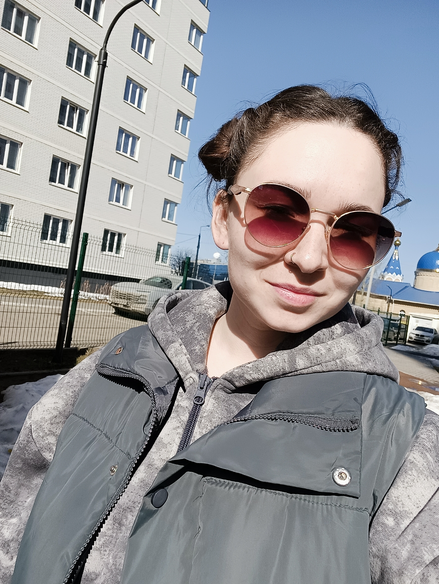 A person with sunglasses and a nose ring is standing outside in Izhevsk, Russia, in front of an apartment building and a blue-domed church. The person is wearing a grey hoodie and a grey vest. Behind them is a fence and a road with a car parked on it. There are patches of snow on the ground. The sky is blue.
