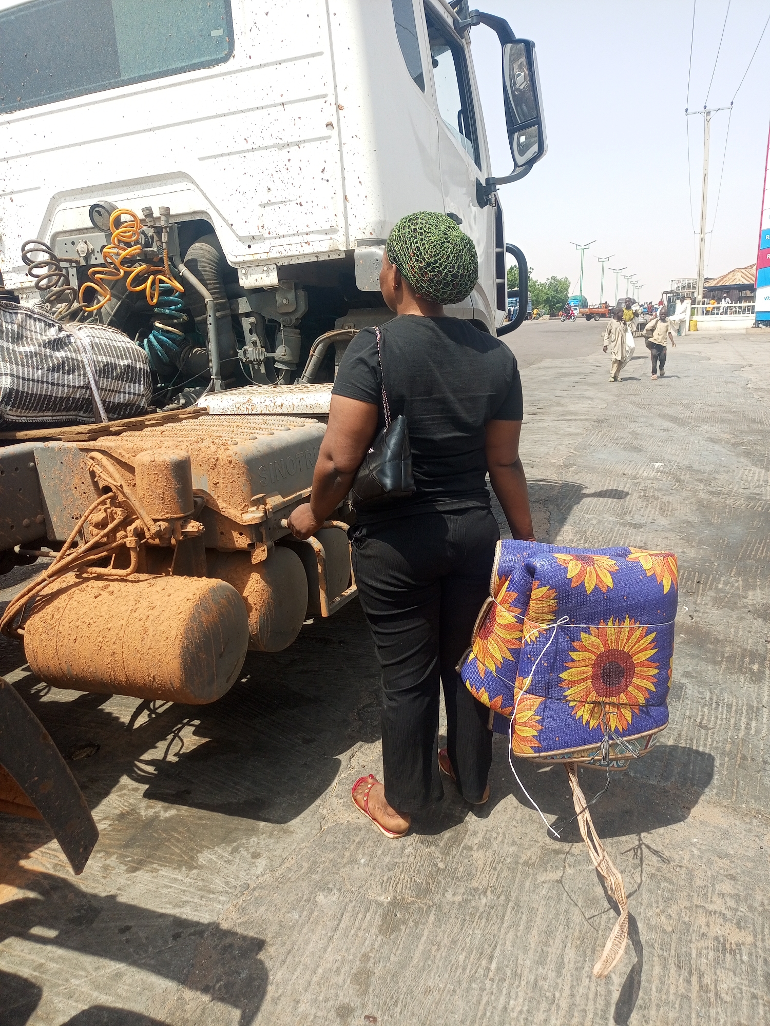 In Gombe, Nigeria, a woman stands beside the dirty chassis of a white truck. She wears a green head covering, black top and pants, and sandals. She holds a blue and yellow floral package and a black purse. The truck's chassis is caked in reddish-brown dirt. In the background, people walk along a street with streetlights, and a building with signage is visible.
