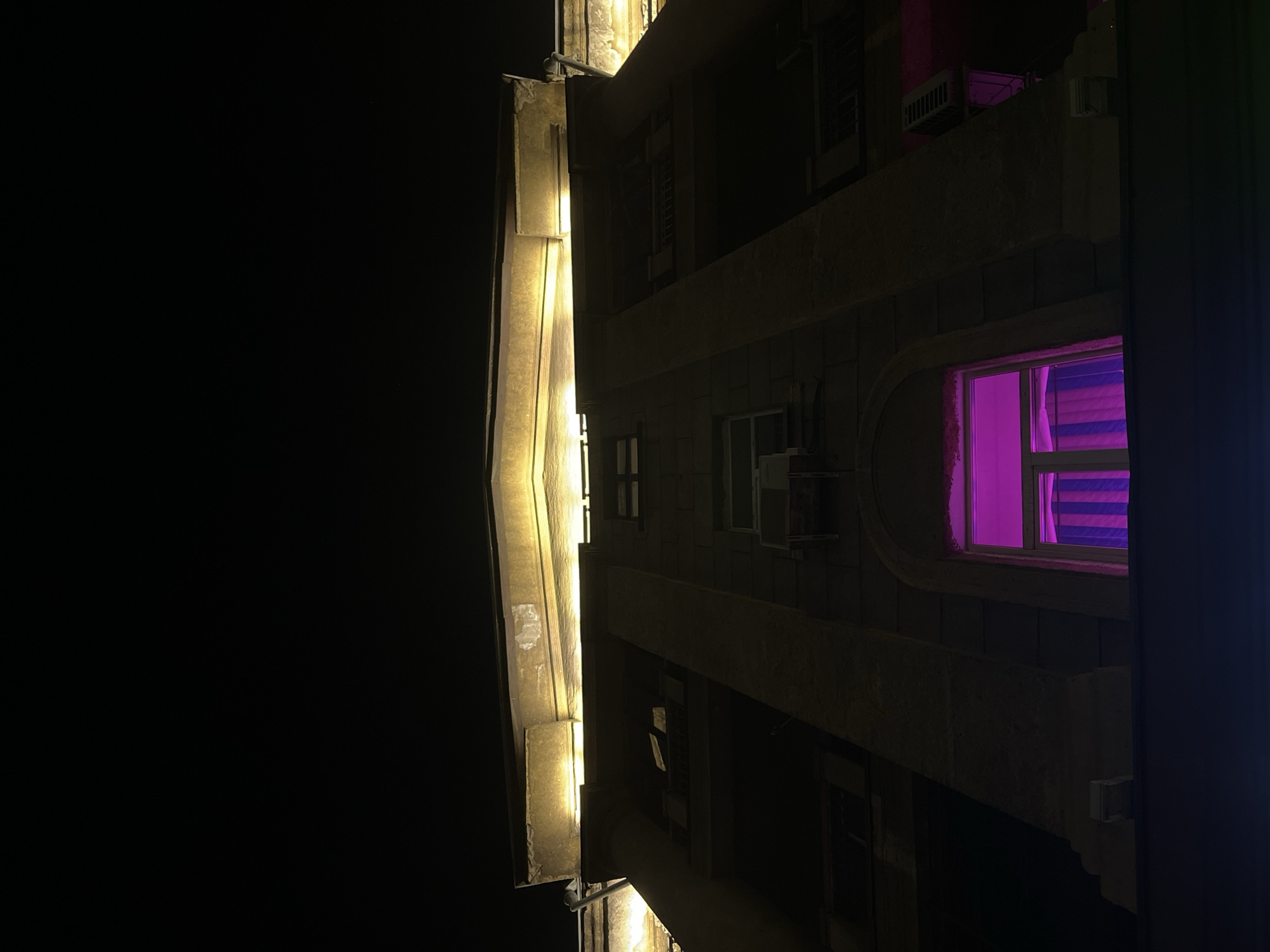 A low-angle shot of a building at night. The building is illuminated by both yellow lights and a pink light emanating from one of the windows. The pink light reflects off the curtains within the window. The rest of the frame is dark, suggesting a nighttime setting. The image suggests that this could be in a urban environment, possibly in Krasnoyarsk, Russia. The building appears to have multiple floors, and there are other windows visible besides the one with the pink light.