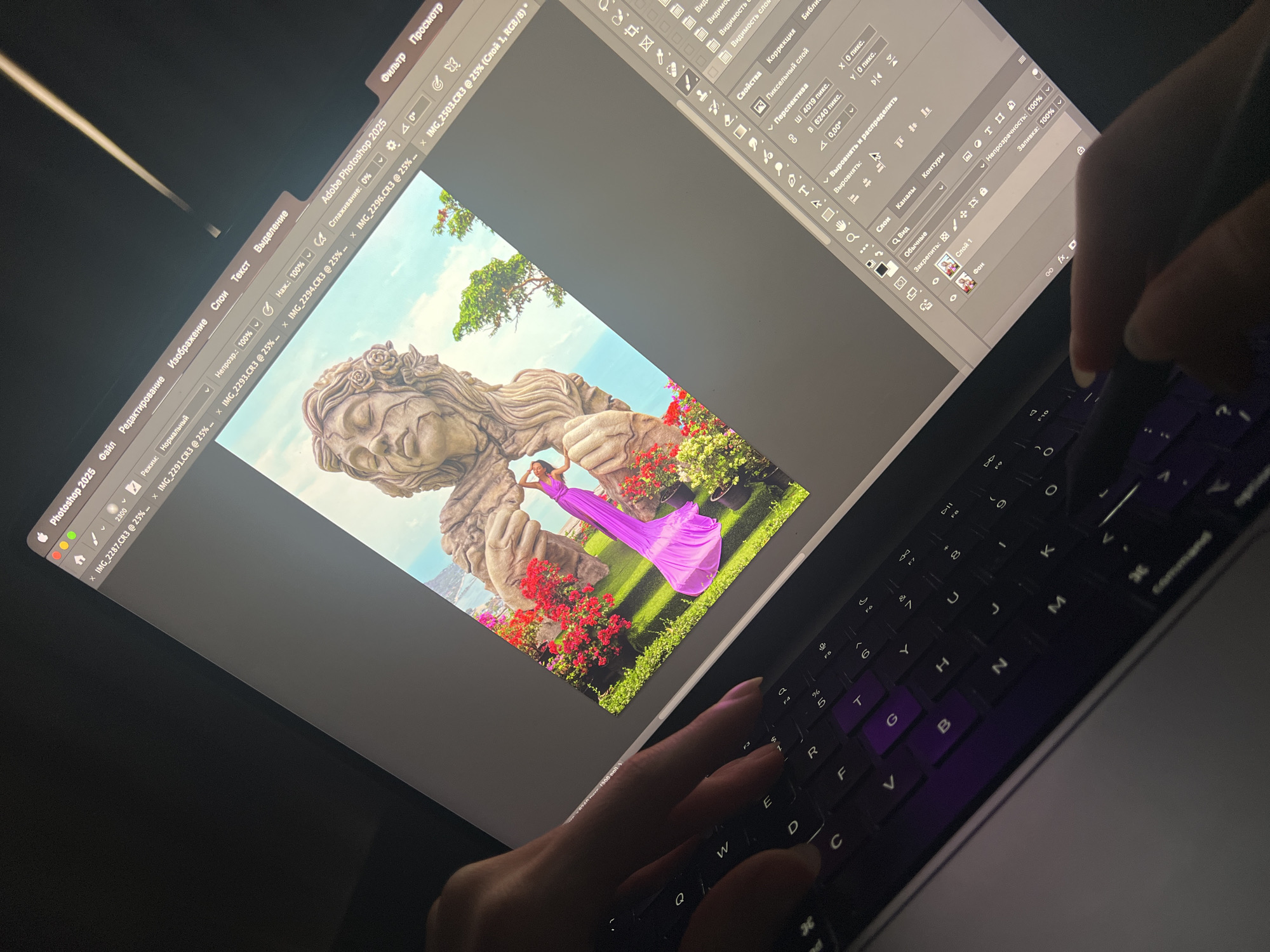 A laptop screen displays the Adobe Photoshop 2025 interface, showcasing an edited image featuring a stone statue and a woman in a long purple dress. Hands are visible, operating the keyboard and using a stylus on the right side of the screen. The application's menus, layers panel, and image editing tools are partially visible. The keyboard has purple backlighting. The location within the city of บ้านบางรักษ์, Thailand is indiscernible from the image.