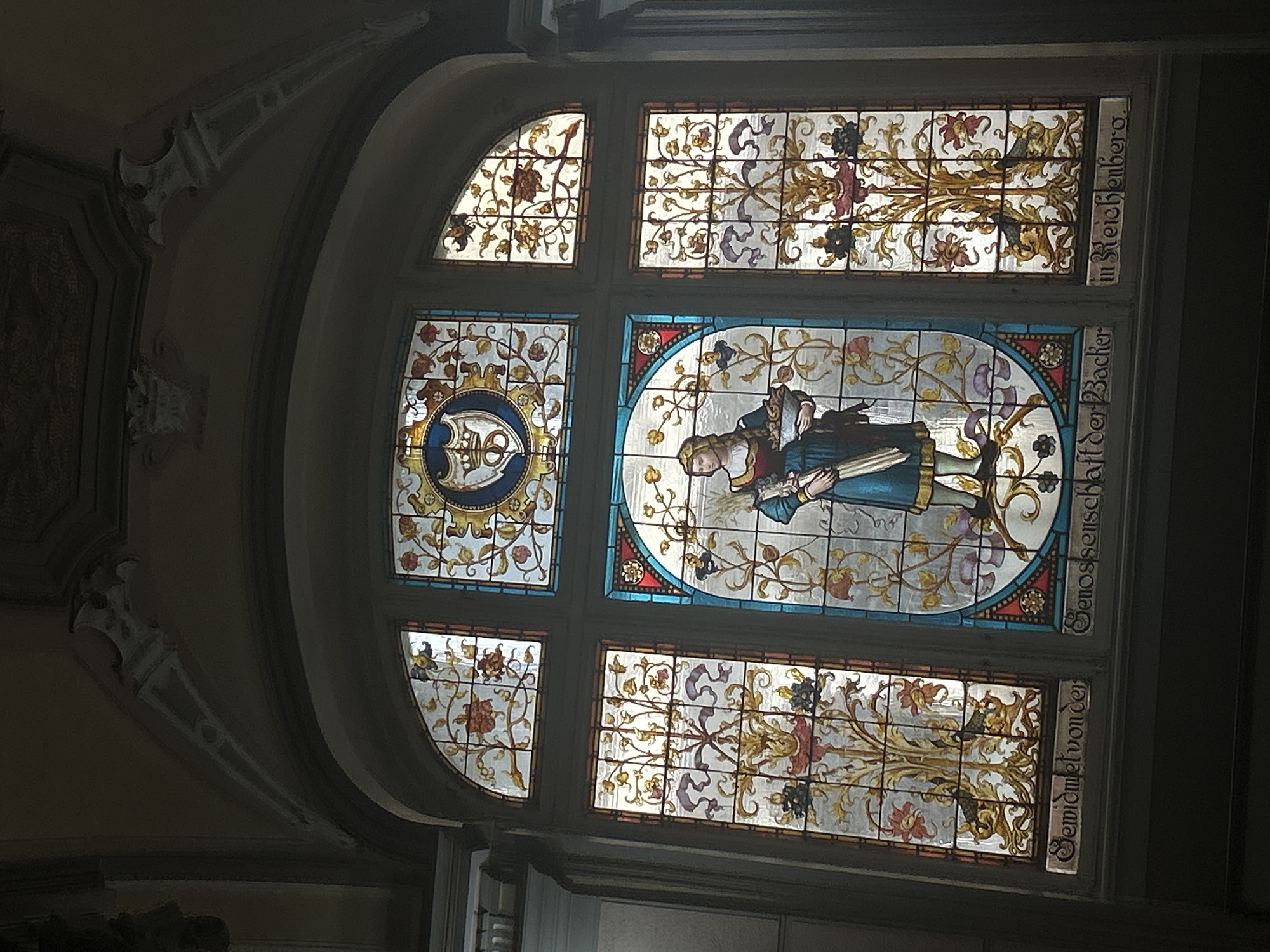 This image showcases a large, elaborate stained glass window within a grand interior setting in Liberec, Czechia. Bright light streams through the window, illuminating its intricate details and vibrant colors.
The window is divided into several sections. The prominent central panel features a female figure, possibly a saint or an allegorical representation, dressed in a blue gown and holding a book or scroll. She is surrounded by delicate floral and foliate patterns in shades of gold, green, and red, bordered by blue and red glass. To her left, another panel contains a circular motif with what appears to be an intricate monogram or symbol, also encircled by ornate floral designs.
Smaller panels above, below, and to the top left of the main sections are filled with decorative floral and organic patterns, primarily in gold, red, and purple against light backgrounds.
The dark frame of the window contrasts with the glowing glass. Text is visible vertically inscribed on the right side of the window frame. The upper inscription reads "in Reichenberg.", identifying the location by its historical German name. Below it, the text reads "Genossenschaft der Bäcker" (Bakers' Cooperative) and "Gewidmet von der" (Dedicated by the), suggesting the window was a gift or commission from a bakers' guild.
The surrounding architecture includes a gracefully curved archway and decorative corbels on the wall, contributing to the historical and ornate ambiance of the space. The scene depicts a richly decorated interior, likely part of a significant building in Liberec. The time of day appears to be daytime, as indicated by the light through the stained glass.