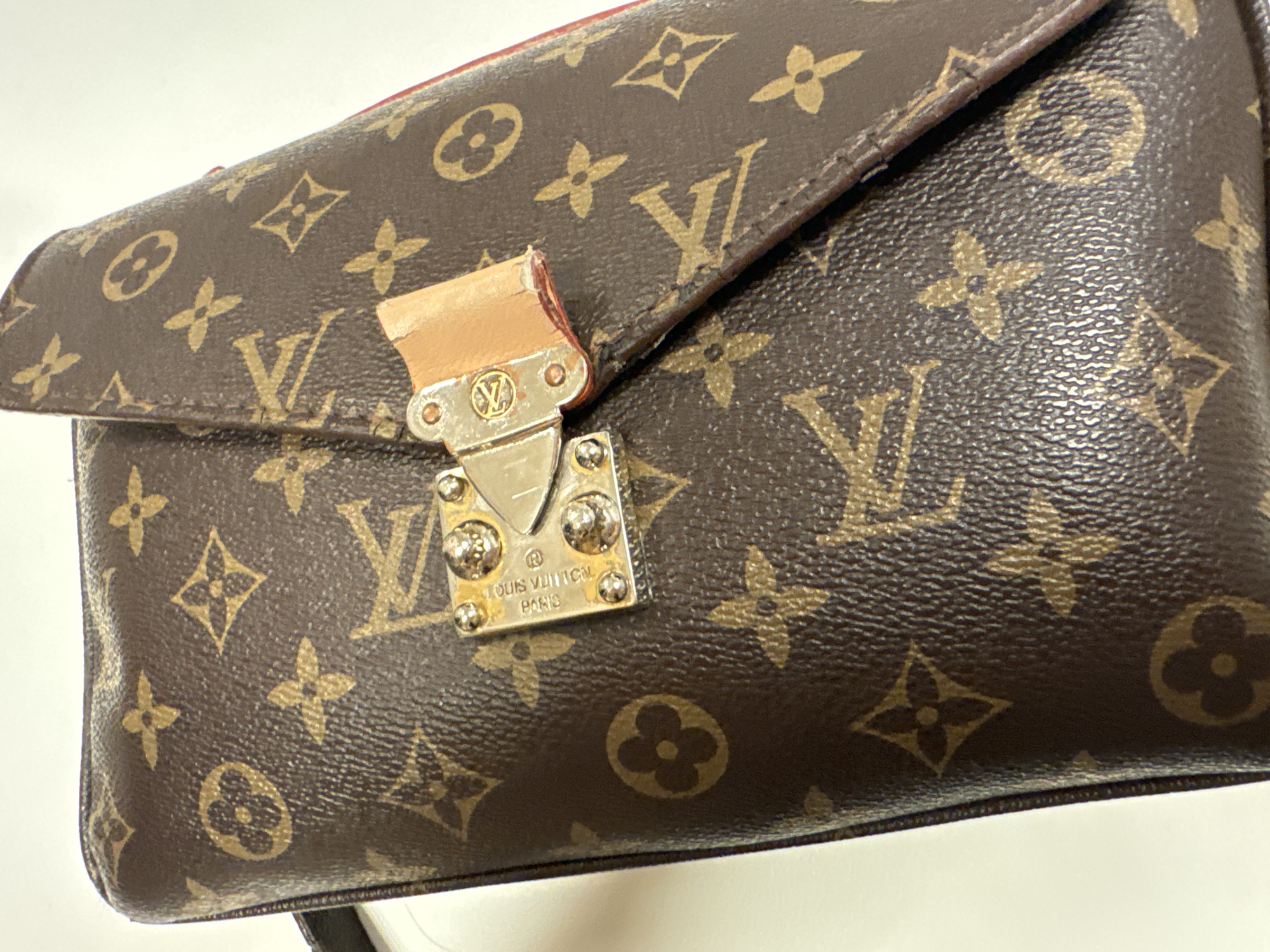 The image shows a Louis Vuitton handbag in the classic brown monogram canvas. The handbag is a flap style with a silver-toned lock mechanism featuring the LV logo and the words "LOUIS VUITTON PARIS" inscribed. The bag appears to be resting on a white surface. No specific location in a city can be determined from the image.