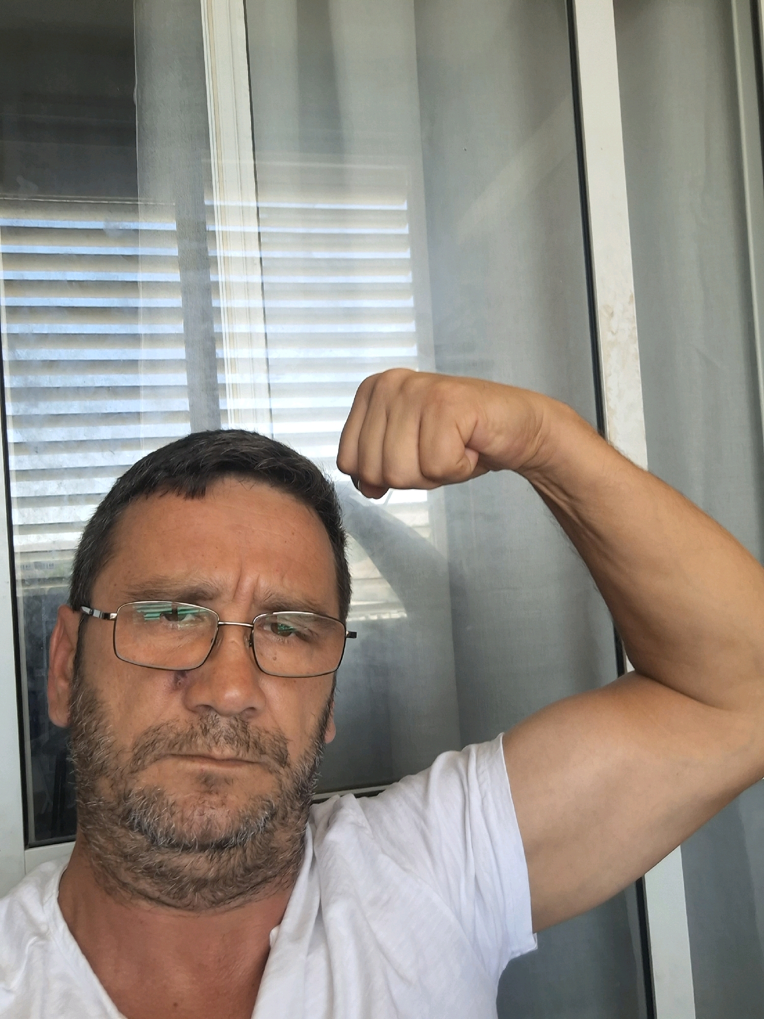 The image is a medium shot of a middle-aged man with short, dark hair, a beard, and glasses, posing with his bicep flexed. He is wearing a white t-shirt. The background appears to be a room with blinds visible through a window. The overall lighting is bright, suggesting it is daytime. There is no identifiable information in the image to indicate its location in Yerocham, Israel.