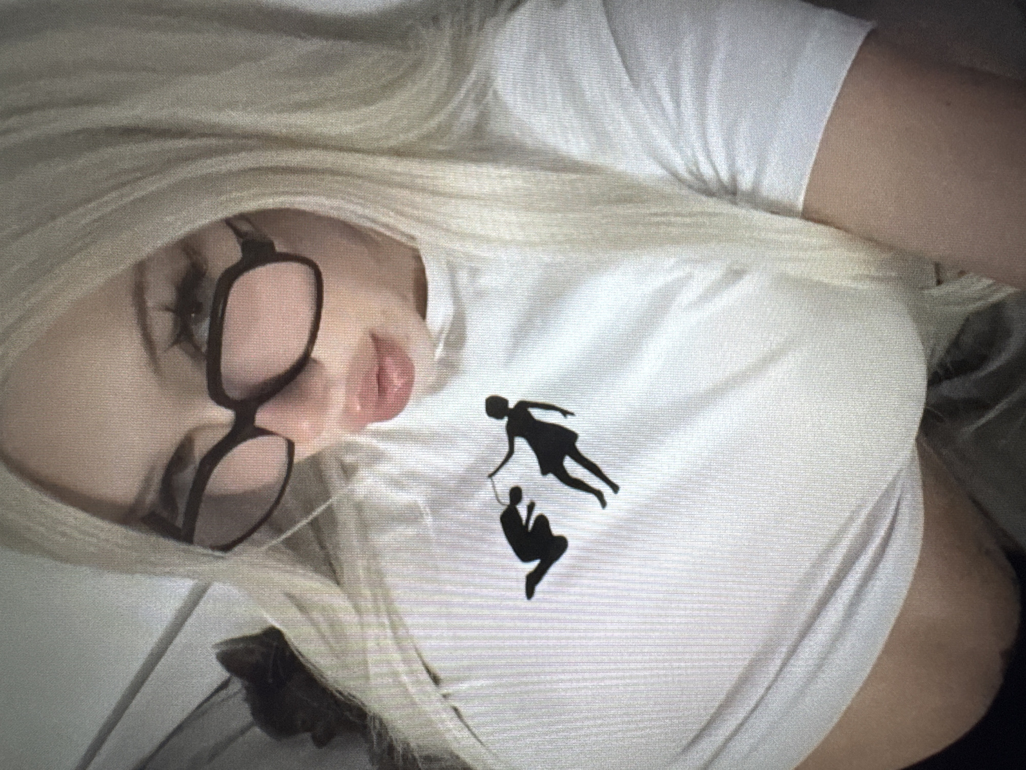 This close-up image features a person, likely female, with long, light blonde hair, wearing black-framed glasses and a white short-sleeved t-shirt. The person is positioned lying down, with their head tilted to the left, and a relaxed expression with lips slightly parted. The background is soft and blurred, showing light-colored fabric, suggesting an indoor setting like a bed or couch.
A notable feature is the graphic design on the white t-shirt, which depicts two black silhouette figures. The upper figure, appearing female with what looks like a dress, is shown floating or flying with one arm extended downwards. The lower figure, appearing male, is depicted in a hanging or swinging posture, holding onto the hand of the upper figure. This design suggests themes of support, interdependence, or a playful connection.
The image itself exhibits a somewhat grainy texture with visible horizontal lines, which might indicate it is a photograph of a digital screen rather than a direct digital capture. There is no discernible text within the image. The specific time of day or precise location cues are not evident beyond it being an indoor scene.