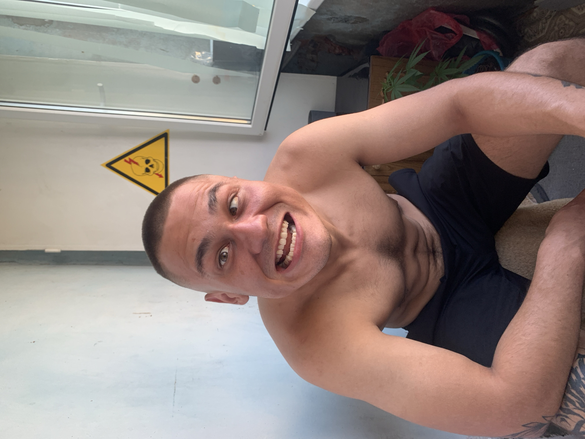 The media file shows a man with a shaved head and a large smile. He is shirtless, wearing black shorts. The image is taken from a low angle, with the man's face dominating the foreground. In the background, there is a window, a yellow triangular warning sign with a skull and lightning bolt, and some plants. The overall impression is casual and informal. The lighting suggests it could be daytime. The presence of a warning sign with a skull and lightning bolt might indicate a hazardous area or a prop for artistic effect. The man's wide smile suggests he is happy or amused. The background elements hint at an indoor or semi-outdoor setting, possibly a greenhouse or a sunroom given the presence of plants. The location context is Pivdennoukrainsk, Ukraine.