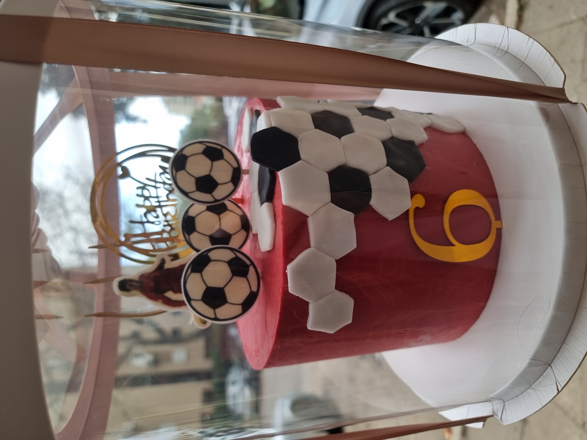 The image shows a round cake decorated with a soccer theme inside a clear plastic box with a tan ribbon. The cake is red with a gold number "9" on its side. Black and white hexagonal fondant pieces are arranged on the top to resemble a soccer ball pattern. Three soccer ball-themed toppers are placed near a "Happy Birthday" decoration. A soccer player figure is also present. The cake sits on a white circular base within the box. The background is blurred, but suggests an outdoor urban environment, likely in Tel-Aviv, Israel, given the mention of this location. A light-colored vehicle is partially visible in the upper background. The photo seems to be taken at an angle, looking down on the cake in the box.