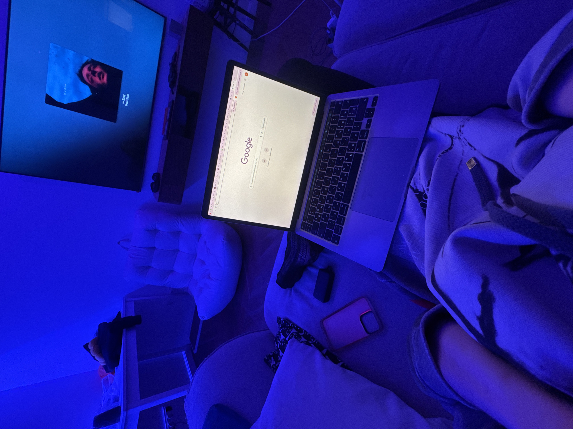 A person sits on a sofa in a room with a blue light. A laptop is on their lap, displaying the Google search engine homepage. A mobile phone and a vaping device rest on the sofa next to the laptop. A TV on the wall displays an image of a person's face. In the background, a white cushion and a small shelving unit are visible. There's no discernible evidence to connect the image to Moscow, Russia.