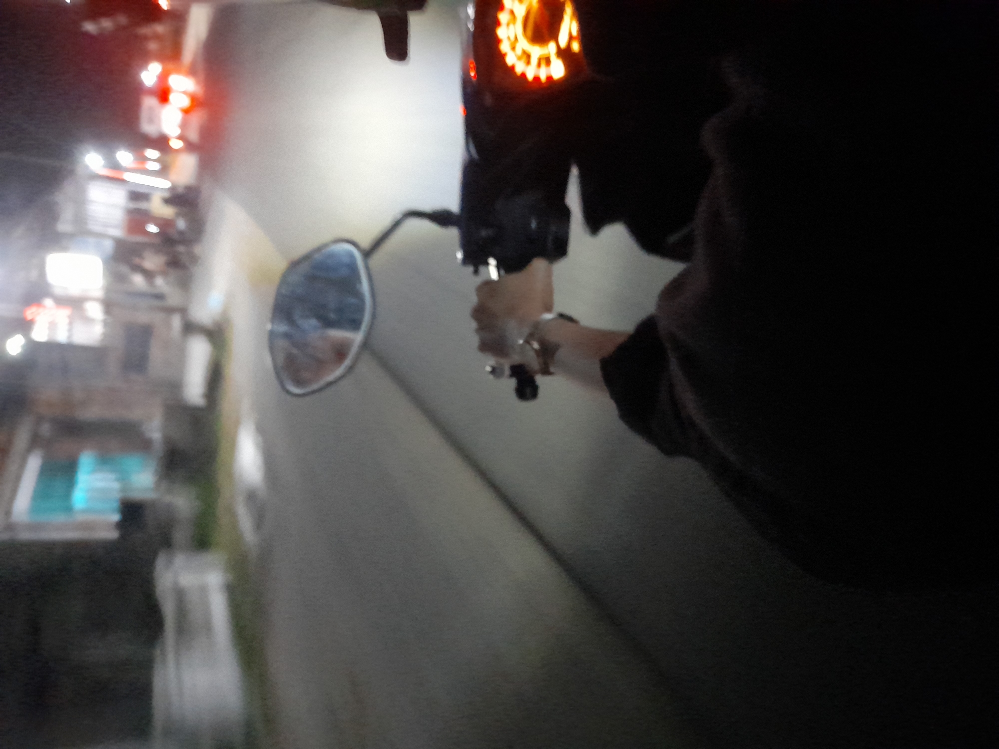The image captures a first-person perspective of someone riding a motorcycle at night on a paved road in Jetaksari, Indonesia. The rider's hands are visible on the handlebars, with one hand gripping the throttle. The motorcycle's dashboard lights up with an orange glow. In the background, streetlights illuminate buildings and other vehicles, including cars with their red taillights visible. The scene suggests movement and travel through a typical urban environment at night.