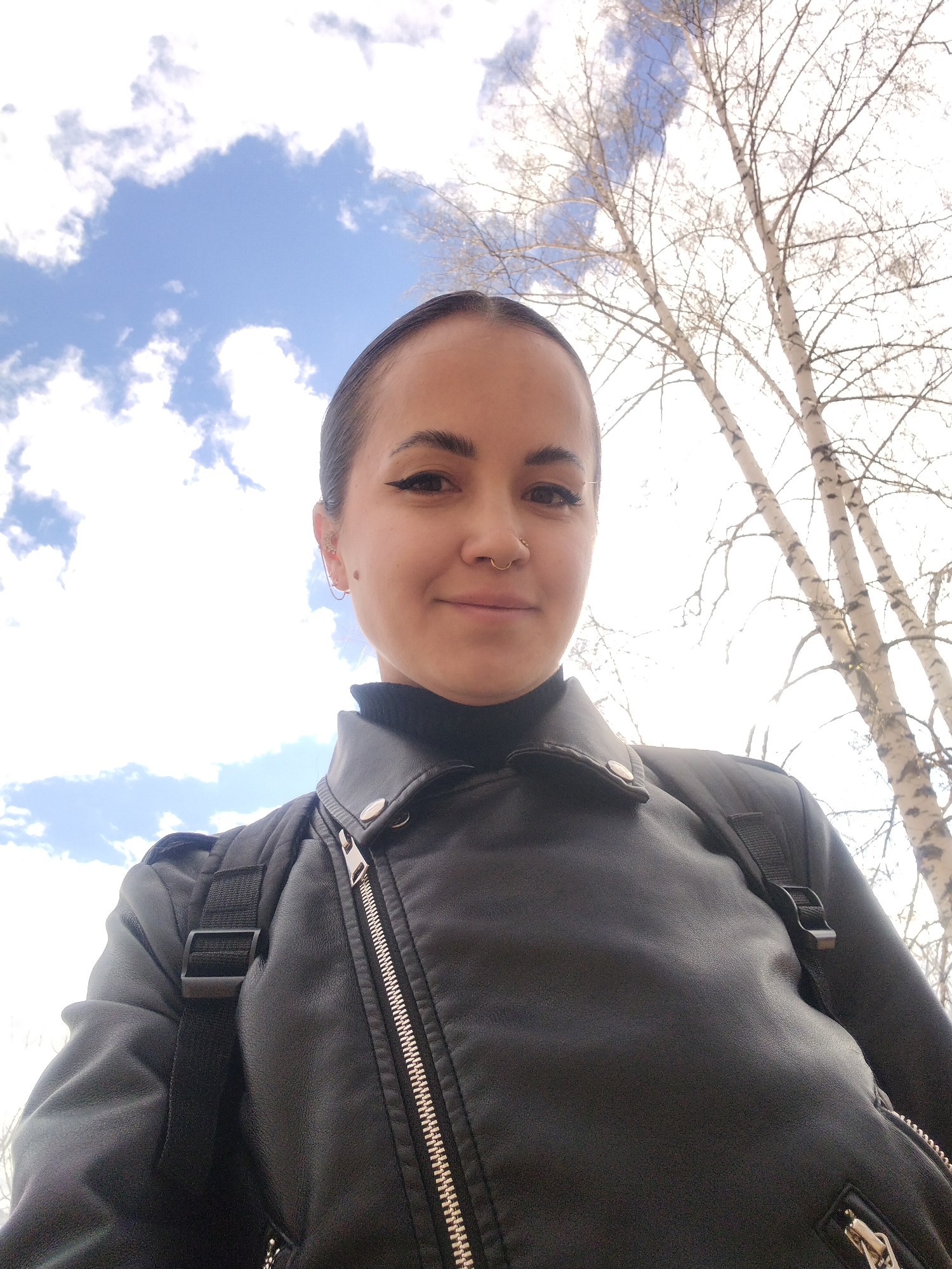 A woman with fair skin and dark hair pulled back is centered in the frame, wearing a black turtleneck sweater and a black leather jacket with a visible zipper. She has a nose ring and multiple earrings on her left ear. The woman is wearing a black backpack.
The background consists of a blue sky with scattered white clouds and tall birch trees with bare branches. The photograph appears to have been taken outdoors during the day. The location is Noviy, Russia.