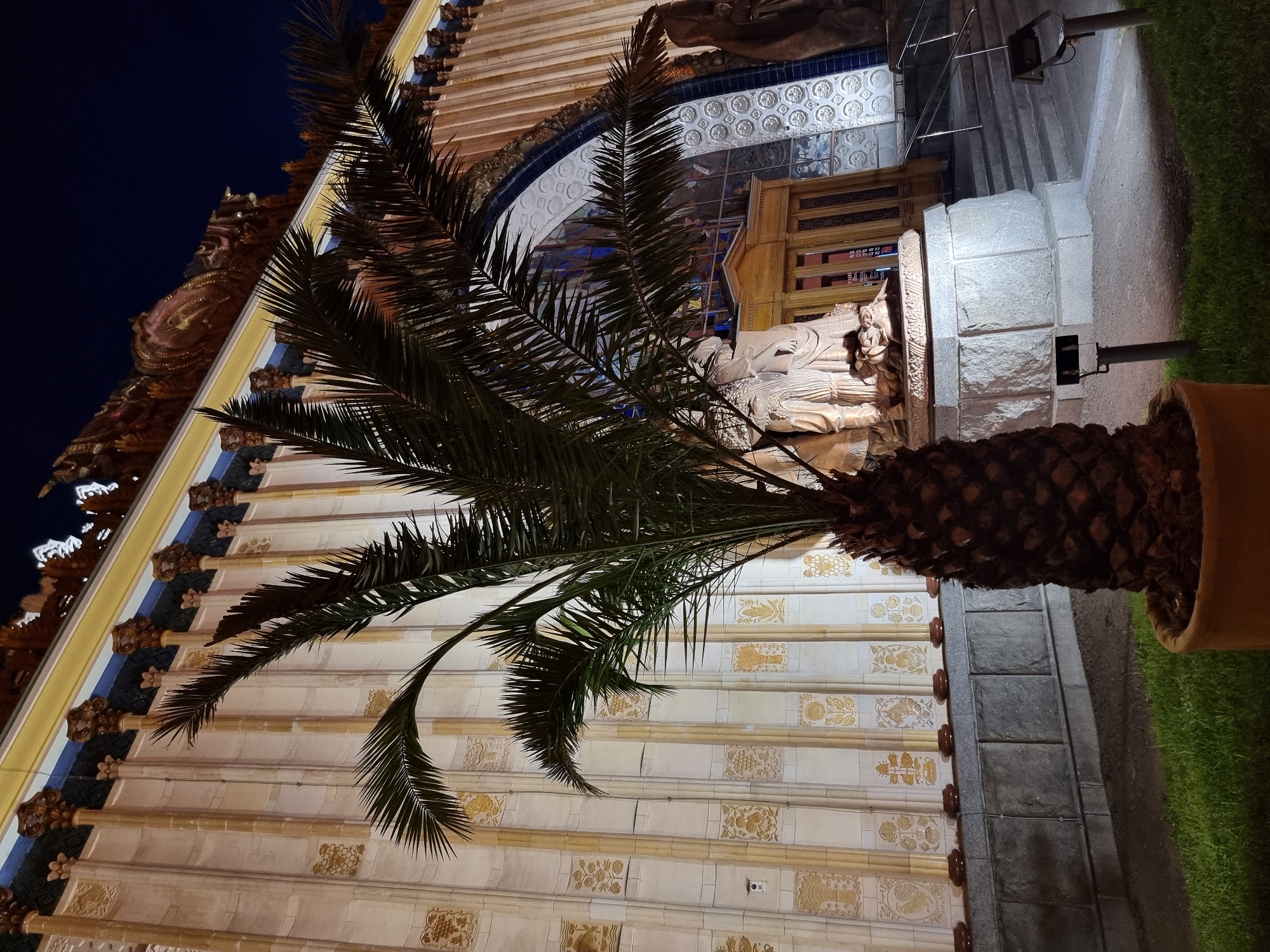 The image shows a palm tree in a pot in front of an ornate building facade at night. The building is lit by artificial lights and features a repeating pattern of vertical fluted columns with decorative capitals. The facade is predominantly cream-colored with gold embellishments. Behind the palm tree, an arched entrance is visible, featuring stained glass windows and a large sculpture of several figures. The area around the building appears to be a plaza or park, with grass and paved areas visible in the foreground. There are also steps leading up to the entrance and what appear to be lighting fixtures. The overall scene suggests a grand, possibly historical or cultural, building in a city setting. Based on the architecture and the presence of palm trees in planters, this location is likely the All-Russian Exhibition Center (VDNKh) in Moscow, Russia, specifically in front of the Ukrainian pavilion, which is known for its distinctive neoclassical and art deco style.