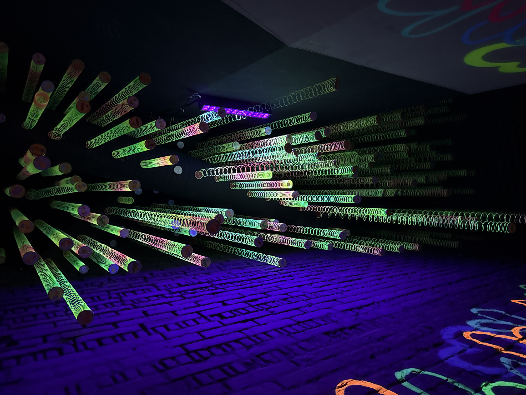 This media file captures an indoor art installation, likely an immersive experience, located in Moscow, Russia. The scene is a dark room illuminated primarily by ultraviolet (UV) or black light, which causes specific materials and projections to glow vibrantly.
The central subjects are numerous Slinky-like springs, predominantly neon green but also displaying hues of pink, orange, and blue. These springs are arranged in dense, multi-layered formations, appearing to project from a dark wall or ceiling on the left and extending diagonally into the depth of the room towards the right. Many springs are attached to or encase cylindrical rods, enhancing their visual structure. Their arrangement creates a dynamic, almost tunnel-like effect, drawing the eye into the space.
The floor is a dark purple-blue, featuring a distinct brick or cobblestone texture that fluoresces under the UV light, emphasizing its pattern. On the right side of the floor, and partially on an upper wall or ceiling section, large, free-form squiggly patterns are projected in glowing orange, blue, and green light, adding to the artistic and playful ambiance.
The overall lighting creates a high-contrast environment where the glowing elements stand out against the deep black background. No people are visible, nor is any text present. The vibrant fluorescent colors and the unique arrangement of objects contribute to a whimsical, futuristic, and slightly psychedelic atmosphere, characteristic of a glow-in-the-dark art exhibit.