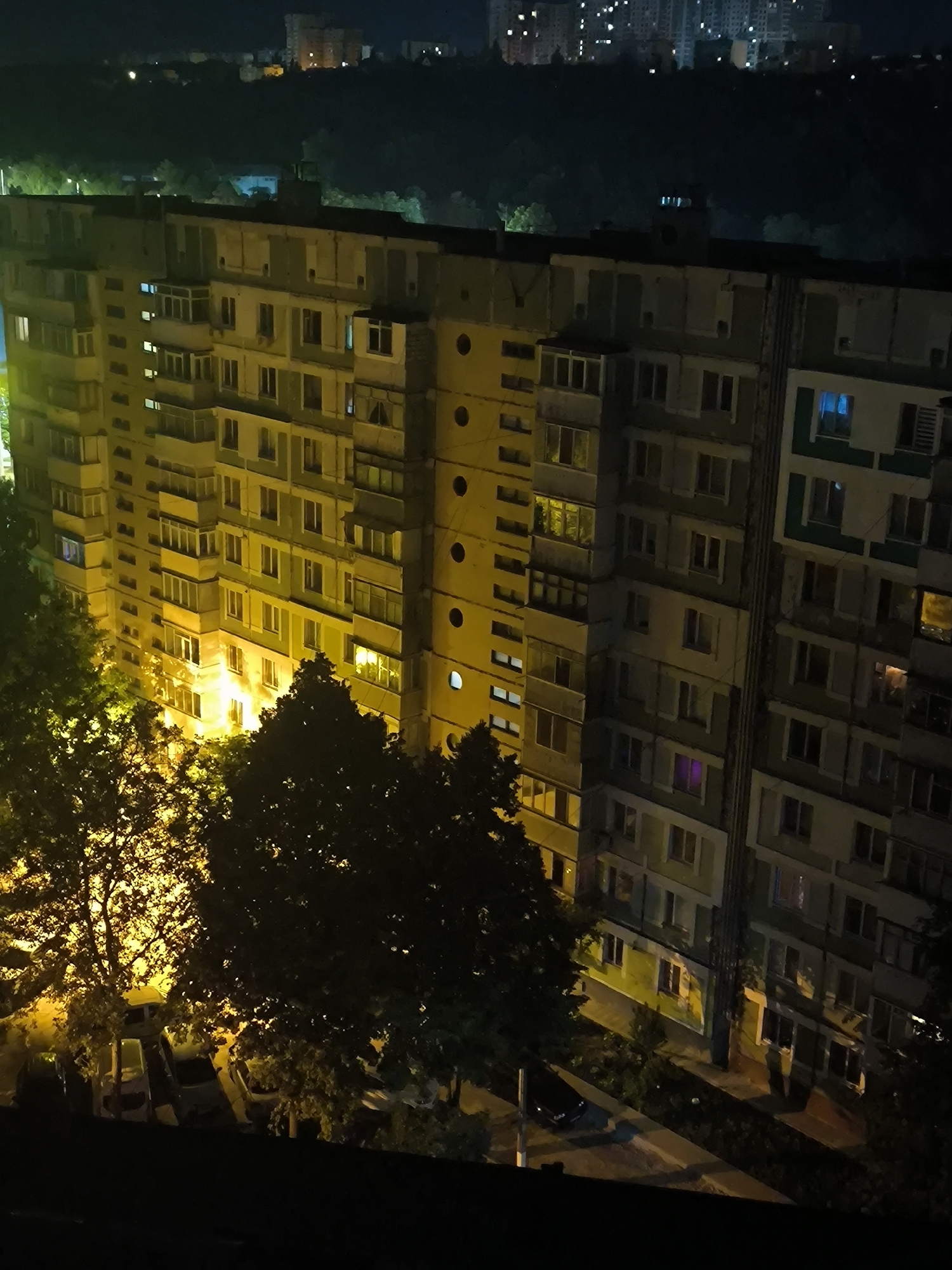 This image shows a night view of a Brutalist apartment building in Chișinău, Moldova. The building is illuminated by streetlights and lights from within the apartments, casting a warm yellow glow on its facade. The building is multi-story, with numerous balconies and windows visible. Trees with dense foliage partially obscure the lower floors of the building, and the branches are also illuminated by the lights. In the foreground, several cars are parked, their forms indistinct in the darkness. In the background, a distant cityscape with more buildings and lights is visible against the dark night sky. The overall impression is of a residential area in a city at night.