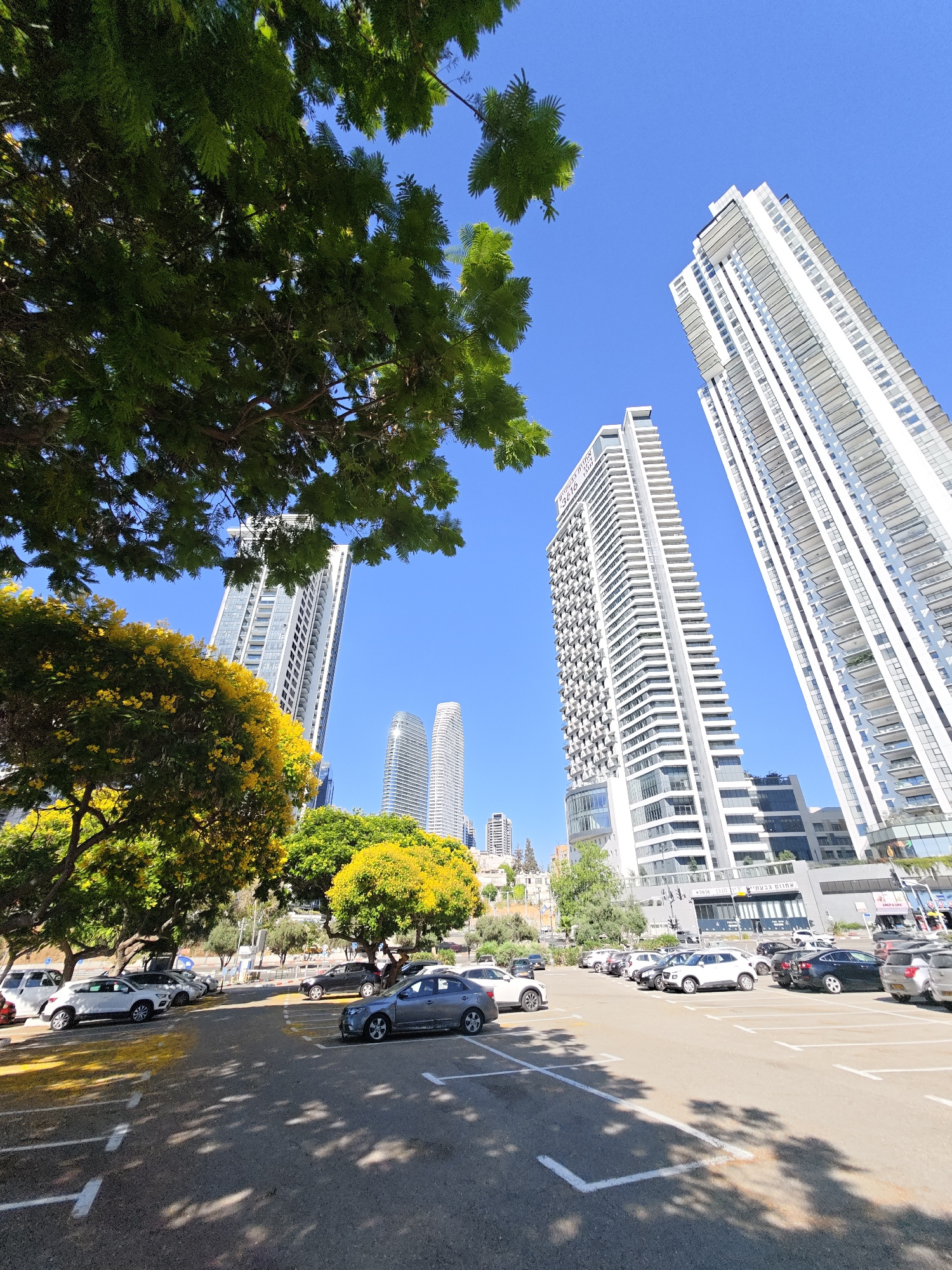 The image captures a bright, sunny day in Tel Aviv, Israel, showcasing a modern urban landscape. In the foreground, a parking lot is visible with several cars parked, including a white SUV and a gray sedan. The ground is asphalt, marked with white parking lines and yellow directional lines. Lush green trees with vibrant yellow flowers frame the scene on the left and in the middle ground, their leaves casting dappled shadows on the pavement.
Dominating the background are several tall, contemporary white buildings. The most prominent are two sleek, high-rise apartment buildings with numerous balconies. Further in the distance, other modern structures and a slightly curved, glass-paneled building can be seen. The sky is a clear, vivid blue. The architecture and the style of the buildings suggest a location in a developed, possibly commercial or residential area of Tel Aviv. The presence of flowering trees adds a touch of natural beauty to the otherwise urban setting.