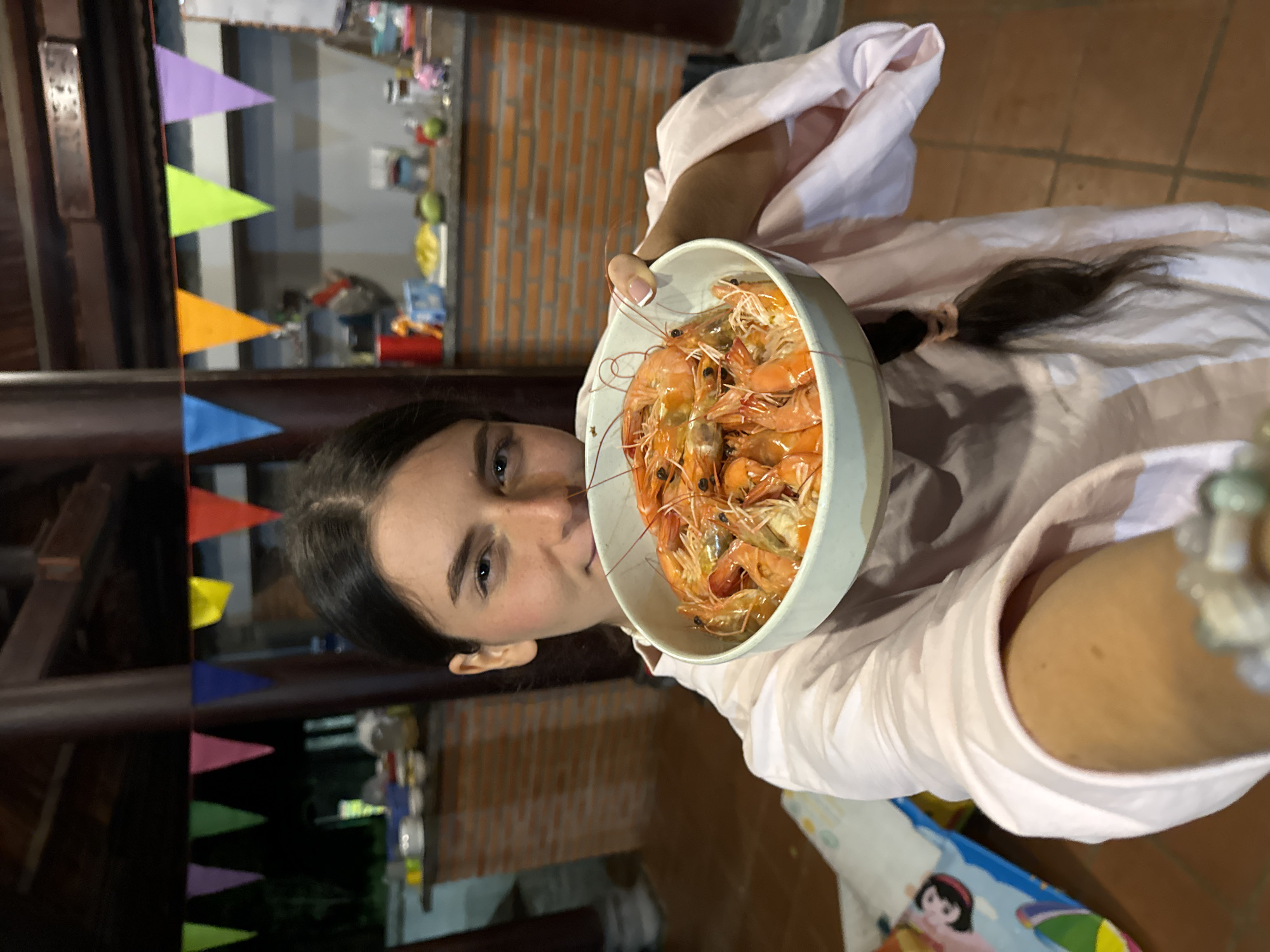 A young woman is prominently featured in a vertical, selfie-style photograph, holding a large white bowl filled with numerous cooked, orange-hued shrimp. She looks directly at the camera with a calm expression. Her dark hair is pulled back in a ponytail, and she wears a light-colored, long-sleeved top, possibly pink or light lavender. Her left hand, visible holding the bowl, has neatly manicured nails with French tips.
The background suggests an evening or night-time setting, with artificial lighting casting a warm glow. A textured brick wall is visible behind her, implying an indoor or semi-outdoor location, characteristic of a casual eatery or a home in Mũi Né, Vietnam. Colorful triangular party flags, or bunting, are strung across the upper background, featuring various shades including purple, lime green, orange, blue, red, and yellow, indicating a festive or celebratory atmosphere. A small portion of a patterned object, possibly a placemat with a cartoon character, can be seen beneath her right arm. The focus of the image is on the woman and the bountiful serving of seafood. There is no discernible text in the image.