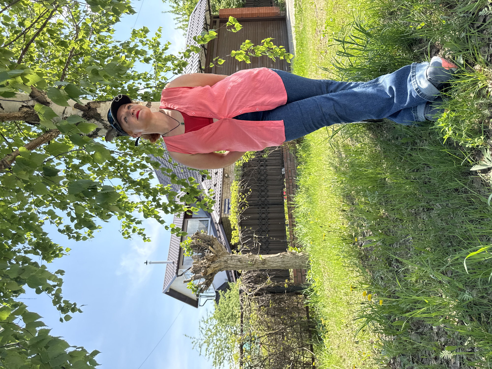 The image shows a woman in Прикамский, Russia, standing in a grassy area near a birch tree. She is wearing a red tank top, a light red open button-down shirt, blue jeans, and sneakers. She also has on a black cap and a necklace. The backdrop includes a residential building with a brown tiled roof and a wooden fence. The sky is partially visible through the tree leaves.
