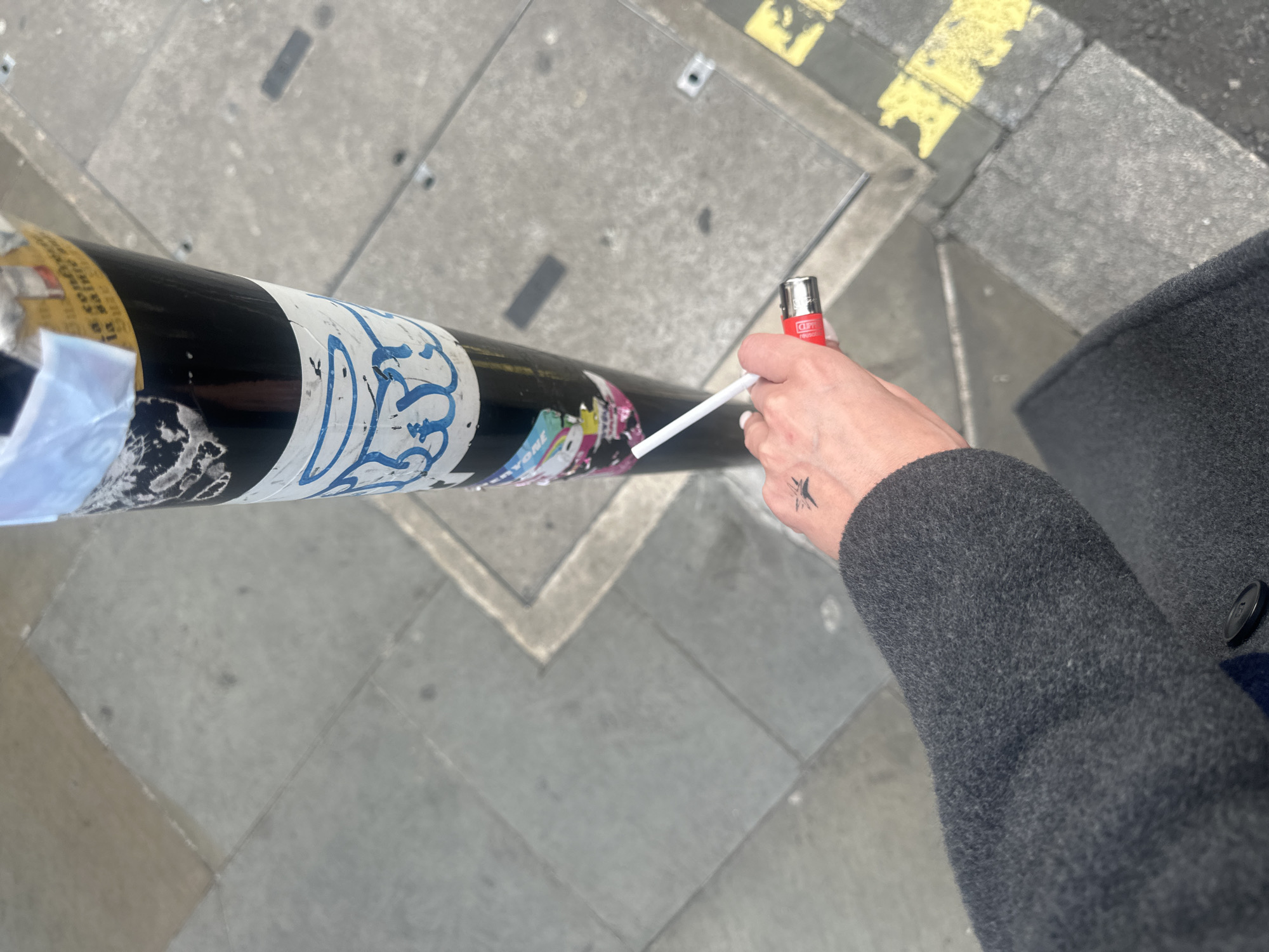 A person stands on a concrete sidewalk next to a black pole covered in stickers, holding a white cigarette in one hand and a red Clipper lighter in the other. The person is wearing a gray coat. The location is possibly in the city of London, United Kingdom. The pole stands near yellow and white painted pavement markings.