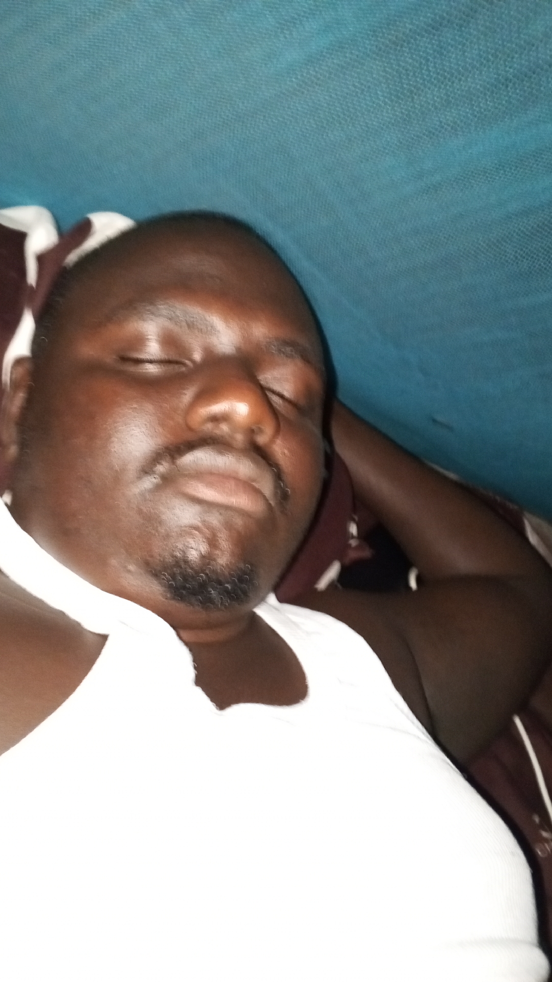 The image shows a man sleeping. He is wearing a white tank top and lying on a bed with a dark brown and white patterned pillow. He has dark skin, a mustache, and a goatee. A blue netting or fabric is draped above him. The location is identified as Unknown, Unknown.
