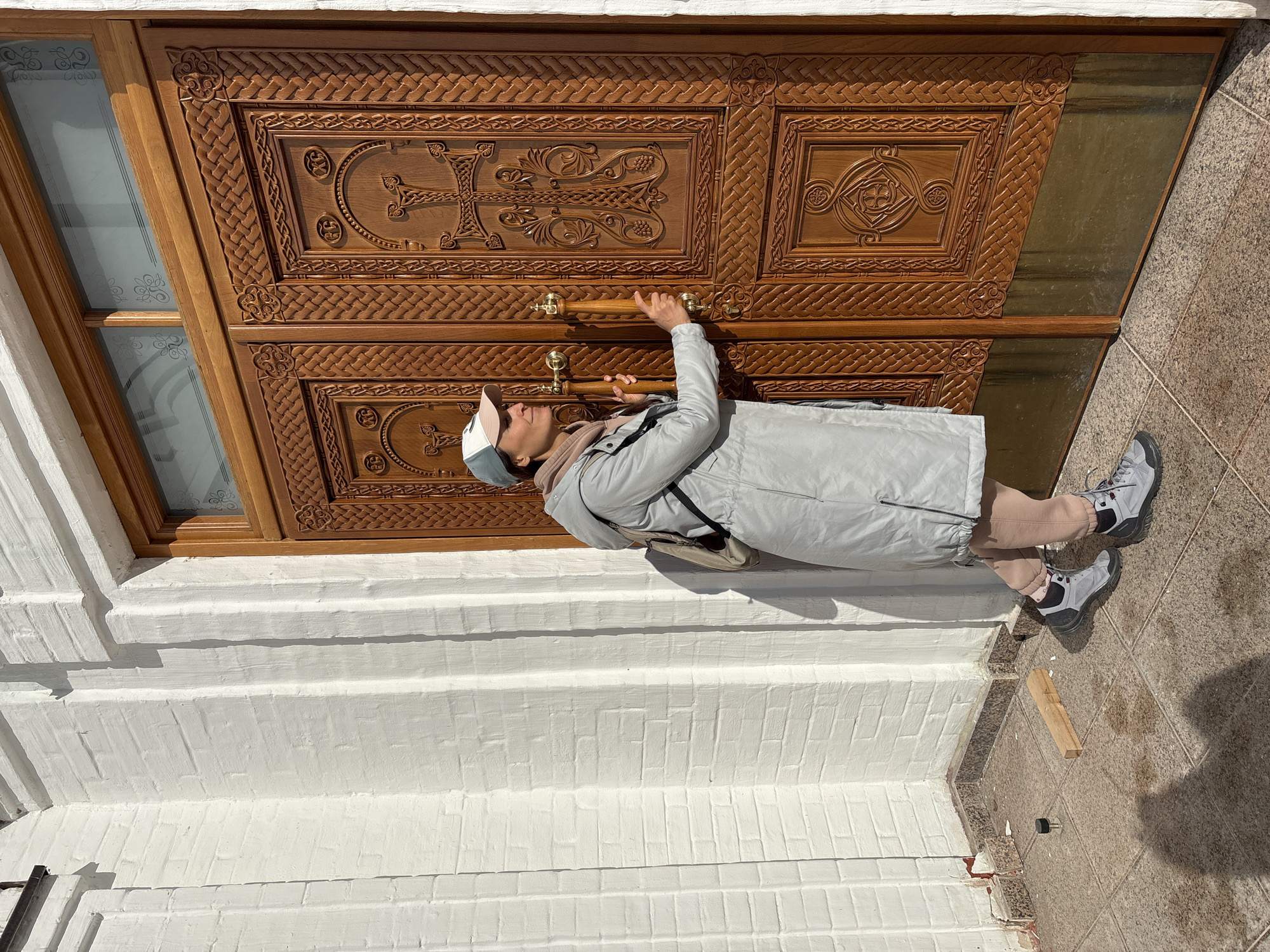 A woman in a gray coat, peach-colored pants, and gray sneakers is pulling on the wooden handles of a large, ornately carved wooden door. The door has two panels with cross-like patterns. The setting appears to be outdoors, near a white brick building. The location is Ulyanovsk, Russia.