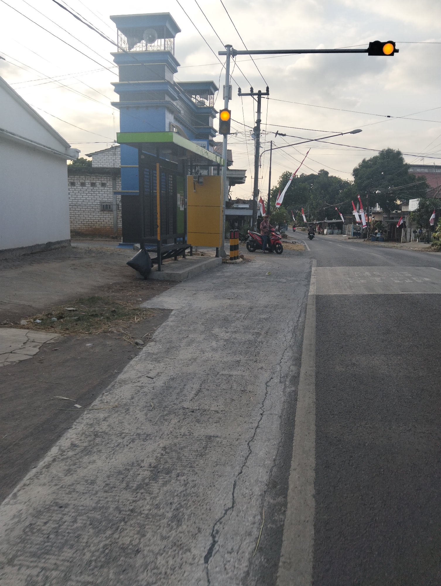 The image depicts a street scene in Dagan, Indonesia, likely near a pedestrian crossing. In the foreground, a grey concrete path transitions to a paved road with a white line marking the lane. To the left, a white building with grey trim is visible, adjacent to a construction of a blue and green structure with a black gate and a yellow kiosk. Several Indonesian flags are displayed along the roadside. A single person is seated on a red motorcycle on the left side of the road, and another motorcycle is in motion further down the street. Traffic lights, with the yellow light illuminated, are present overhead. Power lines crisscross the sky, which is overcast.