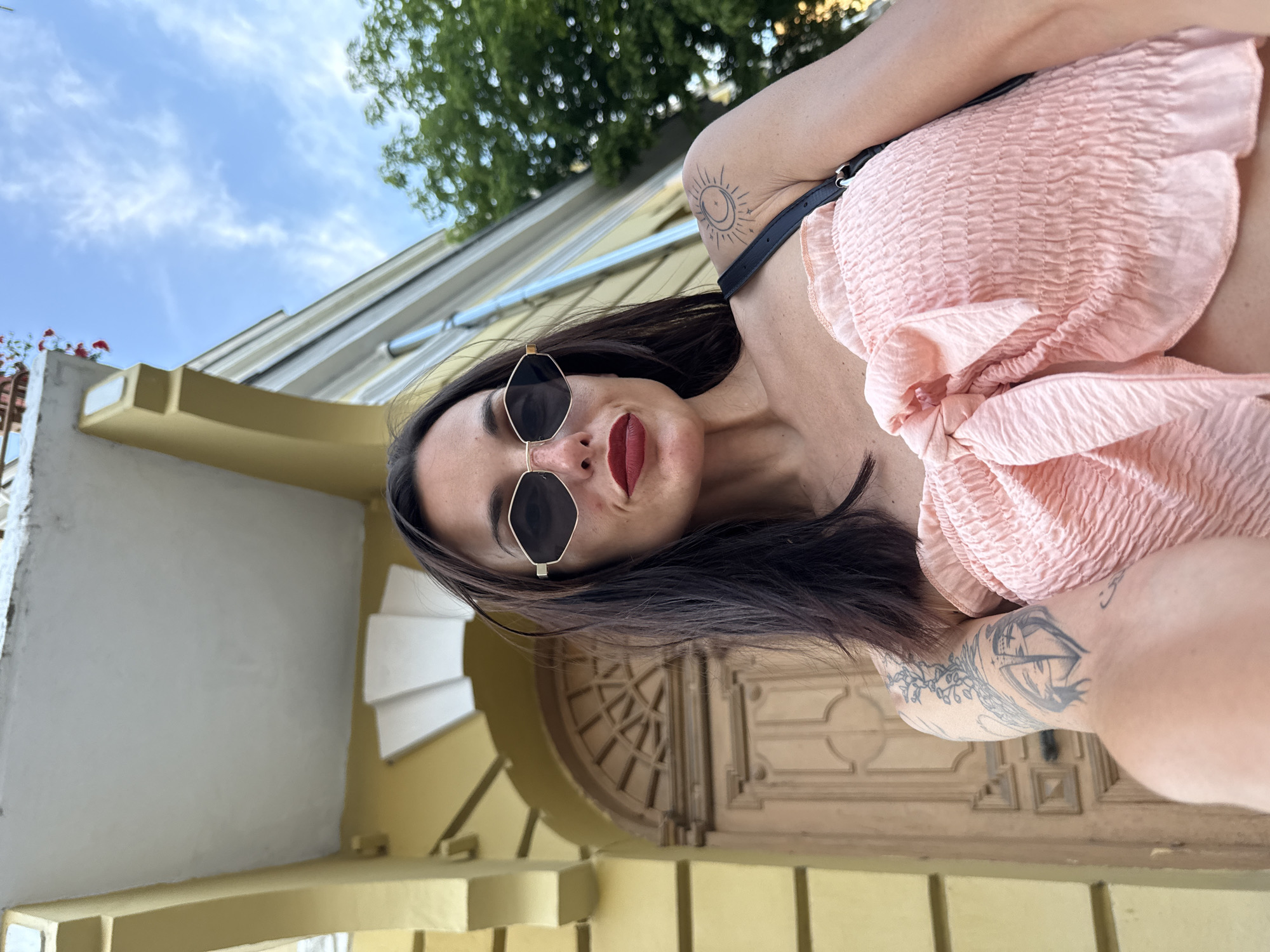 A woman with long dark hair, wearing sunglasses, red lipstick, and a pink smocked top with a bow, poses for a selfie outdoors in front of a yellow building with a wooden door. She has tattoos on her left arm and shoulder. The building appears to be in the city of Pleven, Bulgaria.