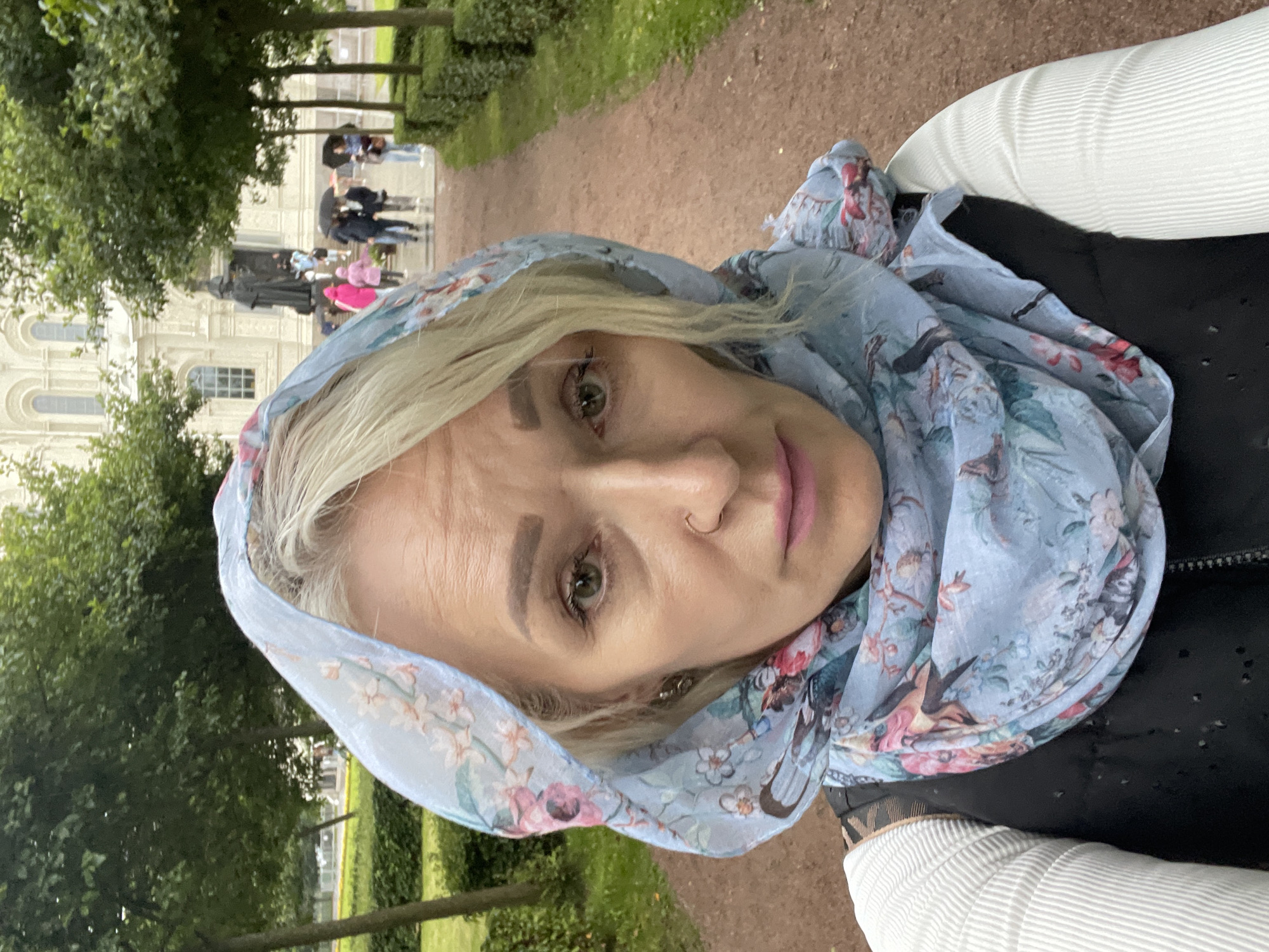 A medium shot selfie taken outdoors shows a woman from the shoulders up. She is wearing a light blue, floral-patterned headscarf, a black vest over a white ribbed shirt, and has blonde hair. Her face is turned towards the camera, with a neutral expression. In the background, a wide, reddish-brown path leads towards a large, ornate white building with arched windows, possibly a cathedral or government building. Several trees with green foliage line the path, and manicured hedges are visible on either side. In the middle ground, a statue of a man on a pedestal is visible, and several people are walking with umbrellas. The environment appears to be a park or a historical site, likely in the city of Kronstadt, Russia, given the architectural style and landscaping. The weather appears overcast, and some of the people in the background are using umbrellas, suggesting it might be raining or has recently rained.
