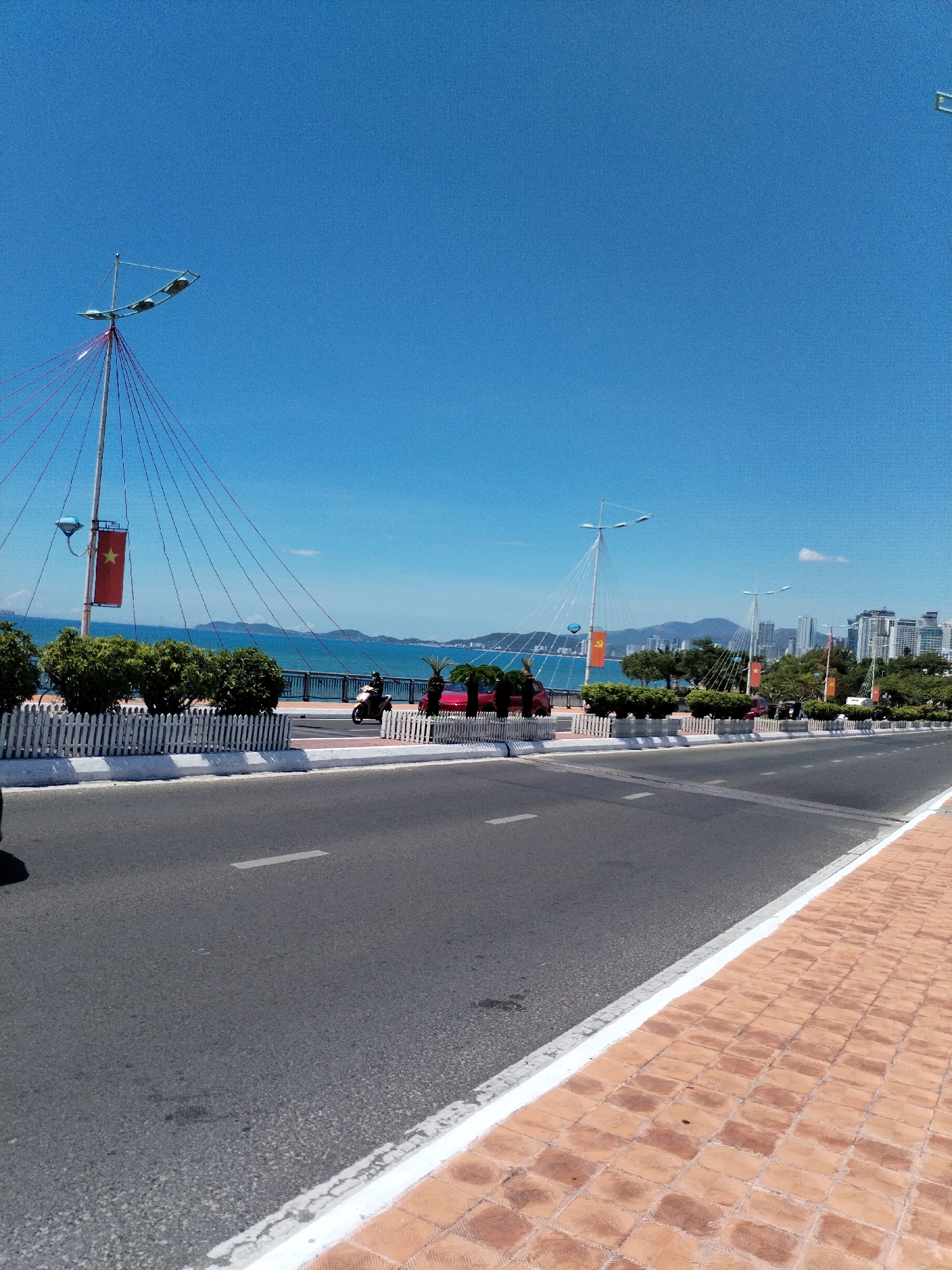 The image depicts a wide, empty asphalt road running parallel to the coast in Bac Nha Trang Ward, Vietnam. On the right side, a red-tiled sidewalk is separated from the road by a white curb. A white picket fence runs along the seaward side of the road, with neatly trimmed bushes and small palm trees behind it. Several tall light poles with red cables and Vietnamese flags are spaced along the coastal road. In the background, the sea stretches to the horizon, with mountains and city buildings visible in the distance. A few vehicles, including a red car and a motorcycle, are visible on the road. The sky is clear and blue.