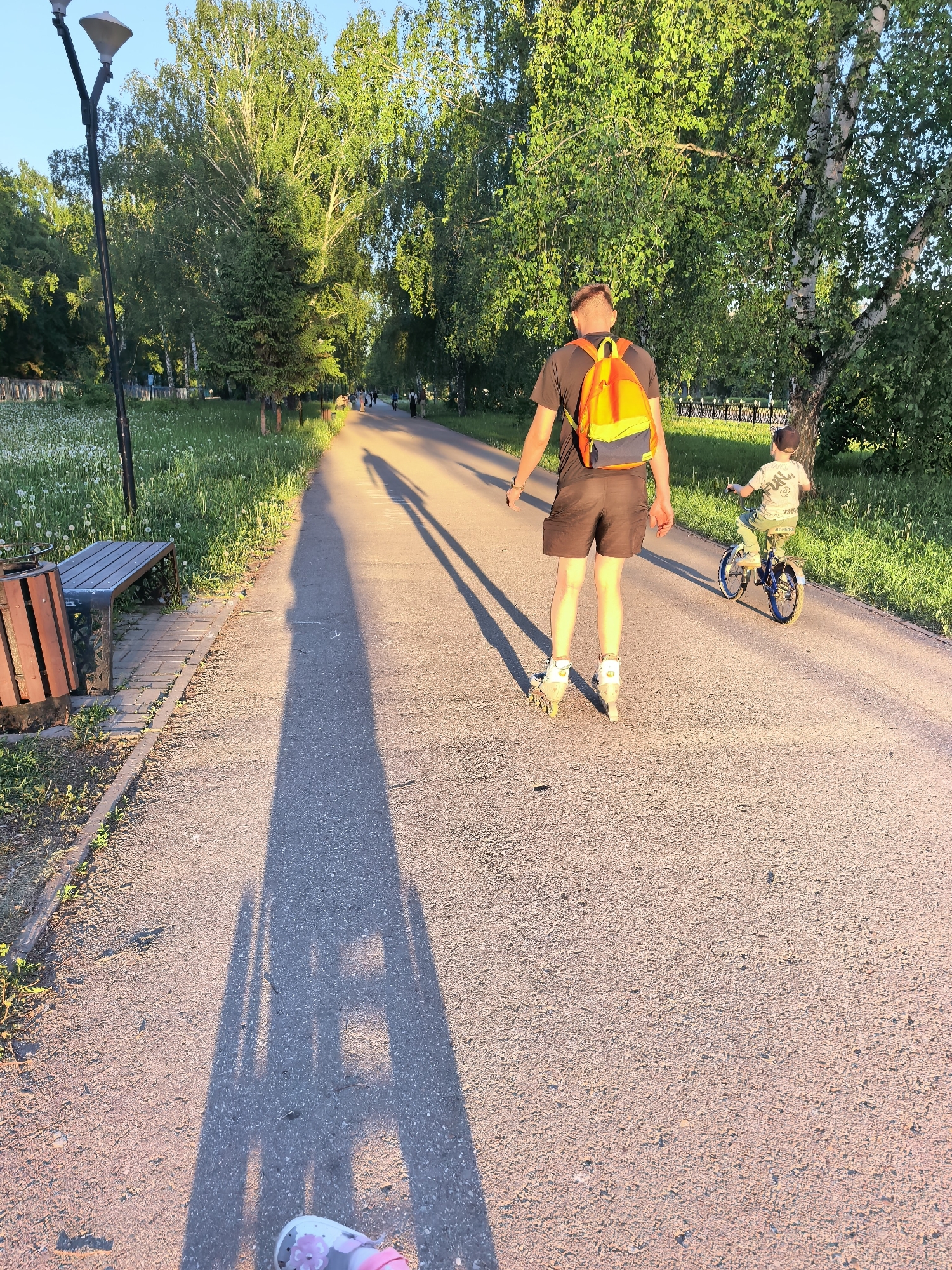 A pathway in a park in Sterlitamak, Russia. A man on roller skates is walking away from the camera, wearing a dark-colored t-shirt, dark shorts, and an orange and blue backpack. A child on a bicycle is slightly behind him and to his right. The shadows of the man and child are cast long on the pathway. A bench sits to the left of the pathway. Trees and green grass line both sides of the pathway. A street lamp stands near the bench. The sky is light blue.
