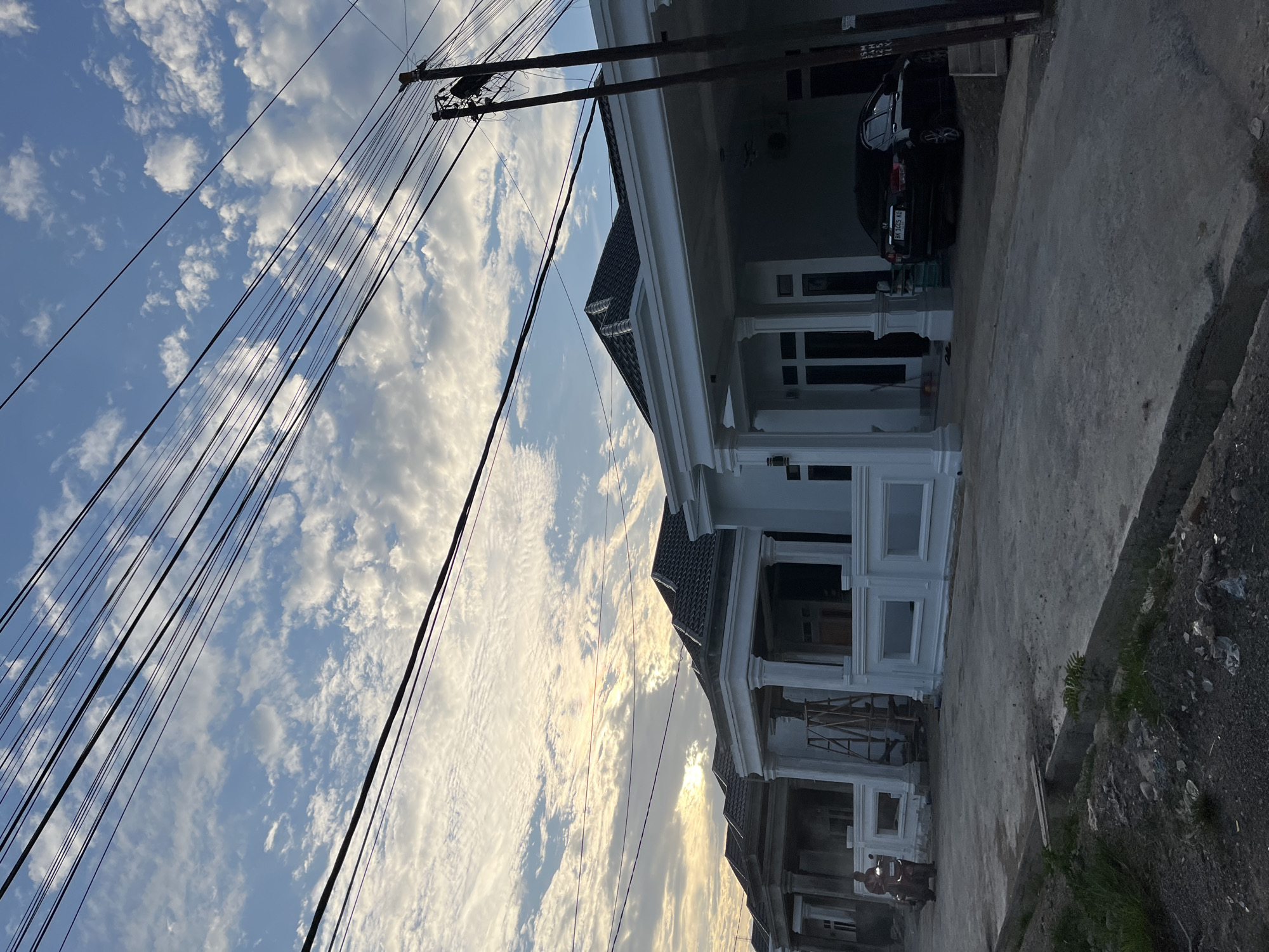 A low-angle, medium shot captures a row of modern, white, single-story houses under a partly cloudy sky. The sky is dominated by streaks of white clouds against a pale blue canvas, with the sun peeking through in the upper left, casting a warm glow. A network of black electrical wires crisscrosses the upper portion of the frame, obscuring some of the sky.
In the foreground, a paved road leads to the houses. To the left, a person on a red motorcycle is riding away from the viewer. The houses themselves feature dark tiled roofs, white facades, and prominent pillars supporting covered porches. One house shows a wooden ladder against its exterior, suggesting ongoing construction or maintenance. A black sedan is parked in the driveway of the house on the right. The environment appears to be a developing residential area, possibly in the city of Lhokseumawe, Indonesia, given the context. The ground in the immediate foreground is rough and unfinished, with a narrow drainage channel. The overall atmosphere is one of calm, late afternoon or early morning light.
