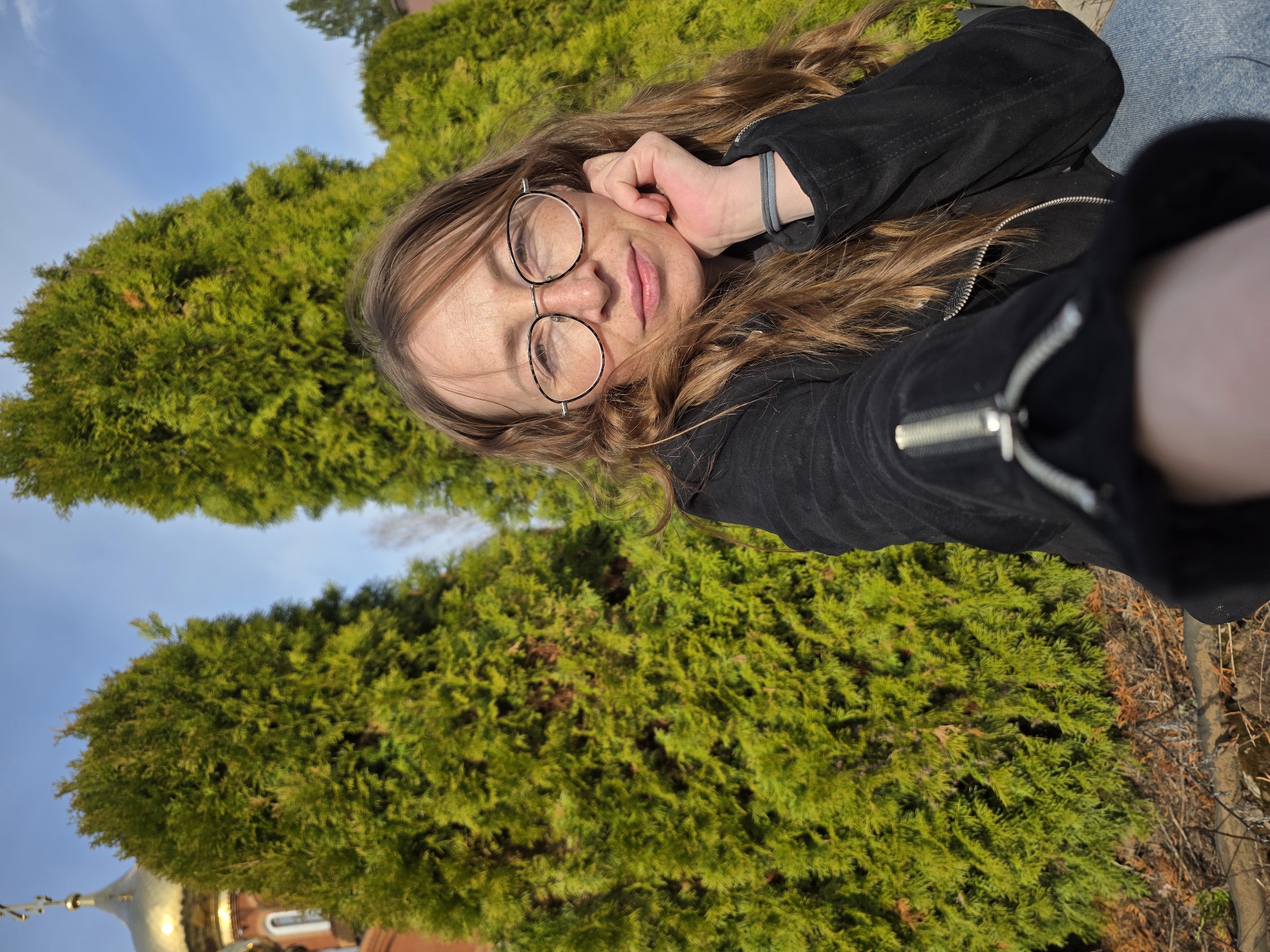 A selfie-style photograph shows a person with light skin, brown hair, and freckles, wearing round glasses. They are reclining and holding their hand near their face. The person is wearing a black jacket with a zipper. Behind them are dense green bushes and trees, with a blue sky visible in the upper left corner. In the lower left corner, the top of a golden domed building is visible. The photograph was taken in the city of Chaikovsky, Russia.