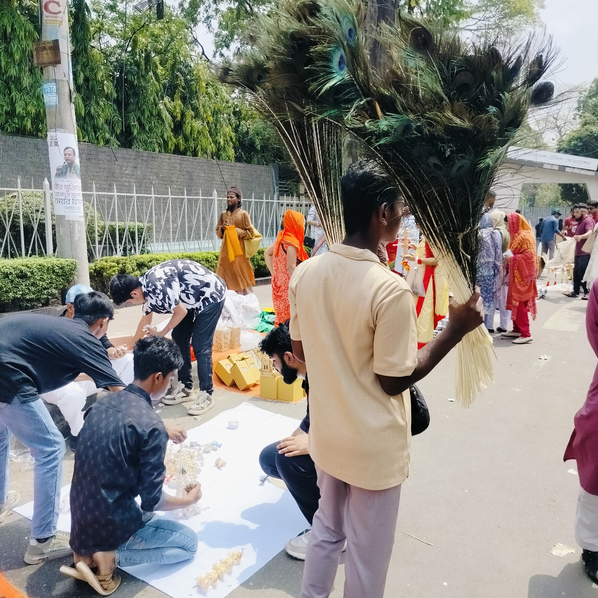 The image depicts an outdoor scene in Dhaka, Bangladesh, likely along a street lined with trees and a low fence. A man stands in the foreground holding a large bouquet of peacock feathers. To the left, a group of men are working on a white sheet spread on the ground, arranging small white and gold objects. Behind them, several people stand in a small gathering, including a man in traditional brown clothing and a woman in an orange sari. In the background, there is a building entrance with the name "বাংলাদেশ টেনিস ফেডারেশন" (Bangladesh Tennis Federation) written above it. A utility pole on the left has posters and advertisements affixed to it, including a poster featuring a man in a green shirt and Bengali script. The overall scene appears to be an outdoor event or preparation for a ceremony.