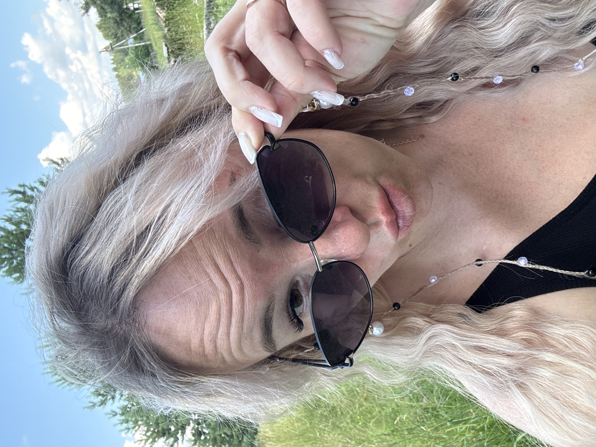 In Pozdyshki, Russia, a close-up, eye-level, selfie-style photograph captures a blonde woman wearing black-rimmed sunglasses. She is outdoors, with a blurred background of green grass and trees under a partly cloudy blue sky. The woman is holding her sunglasses with her right hand, with her index finger and thumb on the frame and temple. Her head is tilted slightly, and she is looking at the camera. She has a playful expression with her lips pursed. Her hair is long and wavy, with some strands falling across her face. She is wearing a black top, and a string with beads and a pearl earring is visible hanging from her sunglasses. Her fingernails are painted white with glitter.
