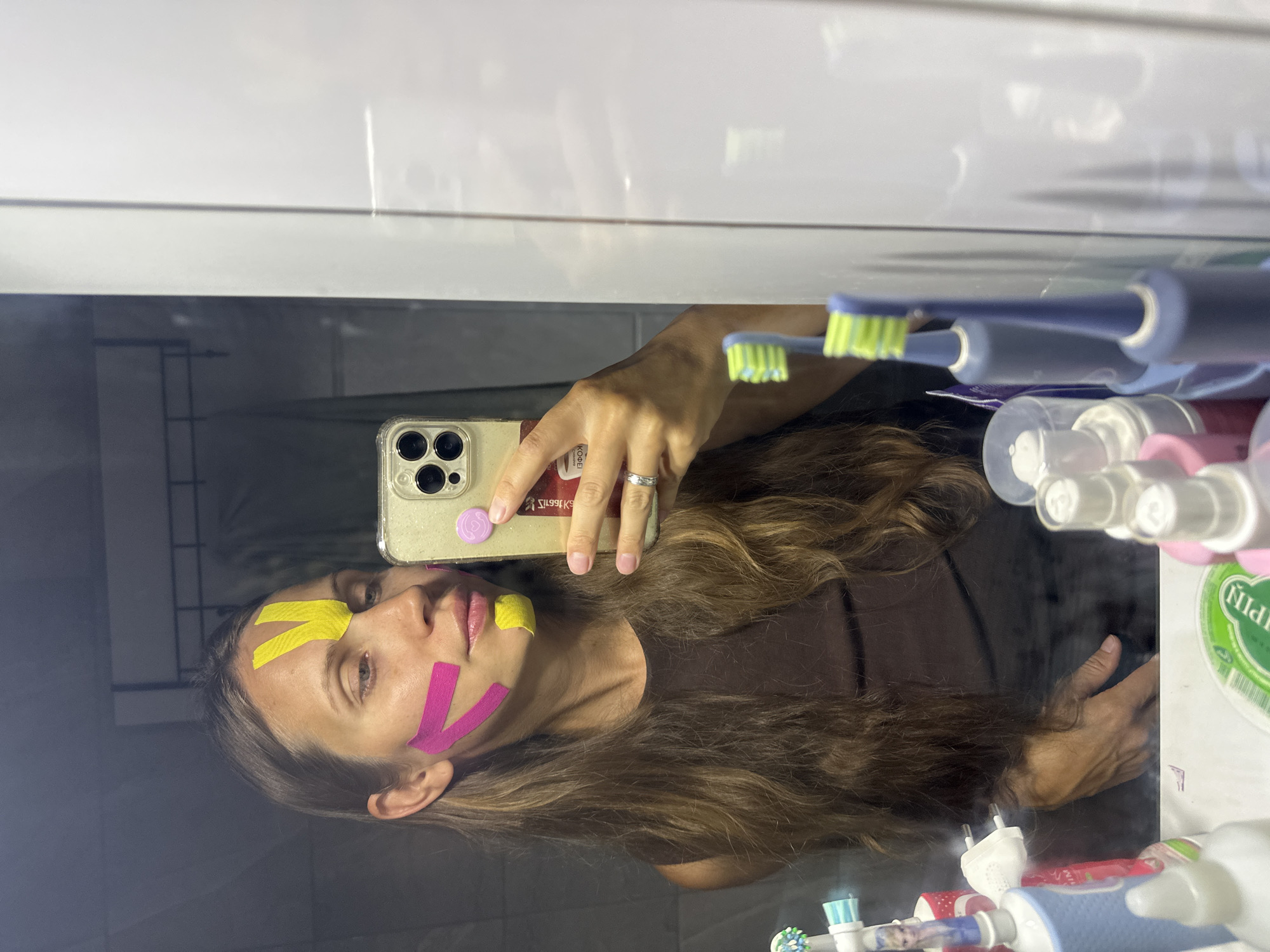 A female individual takes a mirror selfie in a bathroom located in Fethiye, Turkey. The subject has long, wavy brown hair and is wearing a dark, sleeveless top. Her face is adorned with colorful kinesiology tape: two yellow strips form an inverted 'V' on her forehead above the glabella, two pink strips are arranged in a V-shape on her right cheek (viewer's left), and a single yellow strip is placed below her left lip (viewer's right). She holds a silver smartphone, featuring a triple camera system and a clear protective case, in her right hand. A pink circular phone grip with a lighter pink swirl design is affixed to the phone's back, along with a sticker displaying text including "КОФЕ" (Russian for "coffee"). A silver ring is visible on her left ring finger, and her fingernails are painted light pink. The mirror reflects a bathroom counter below, which holds several items: two blue electric toothbrushes (one with a character design, possibly Elsa from Frozen), an additional blue toothbrush head, and various bottles including a pink spray bottle and a white squeeze bottle. Part of a green-labeled container and a white electrical plug are also visible on the counter. The background reflection behind the woman shows a darker room, possibly a shower area, with a rack or rail visible. To the right of the mirror, a clean white wall is reflected.