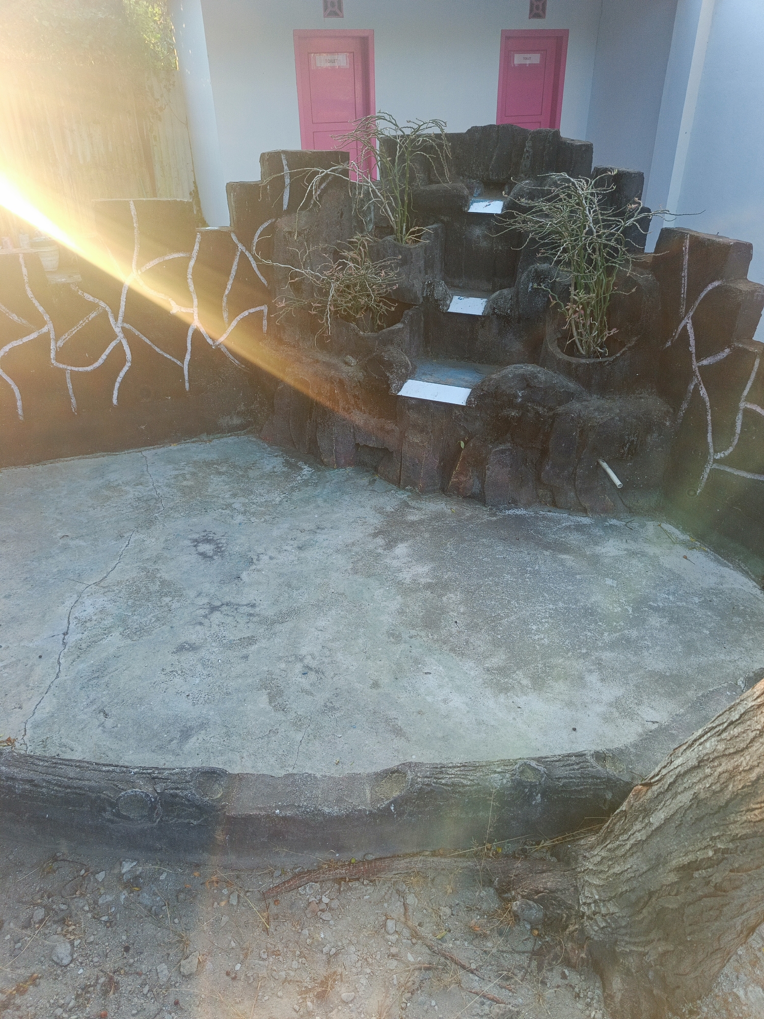 The image depicts an outdoor scene with a concrete basin in the foreground, suggestive of a dry fountain or pond. A decorative rock-like structure with multiple levels is built into the back of the basin, with sparse, dry-looking plants growing in pots on its ledges. The structure is painted with white, abstract lines. In the background, a light-colored wall is visible with two pink doors, each labeled "TOILET" and topped with a decorative emblem. The sun is shining brightly from the left, casting strong lens flares across the image and illuminating the scene with a warm, golden light. The ground around the basin is covered in dirt and gravel, with visible tree roots and fallen branches. The location is described as being in the city of Gorontalo, Indonesia.