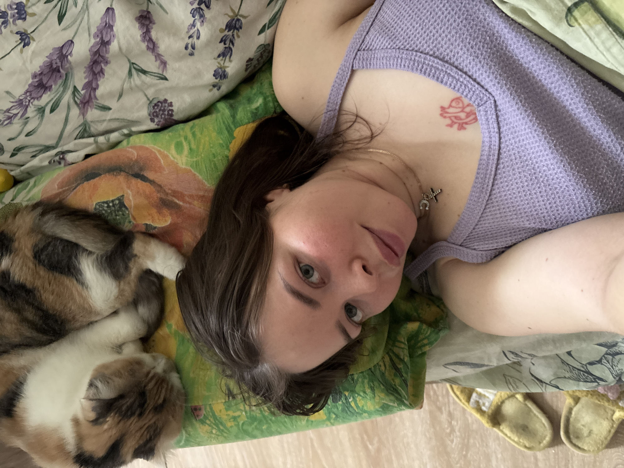 A woman in a lavender tank top lies on patterned bedding with a calico cat. The woman has dark hair with some gray streaks and light eyes. She wears a gold chain with a cross and horseshoe pendant. A red tattoo is visible on her chest. The bedding includes a floral patterned pillow and green and floral patterned sheets. Yellow slippers are visible in the background. The location in Izhevsk, Russia cannot be determined based on this image.