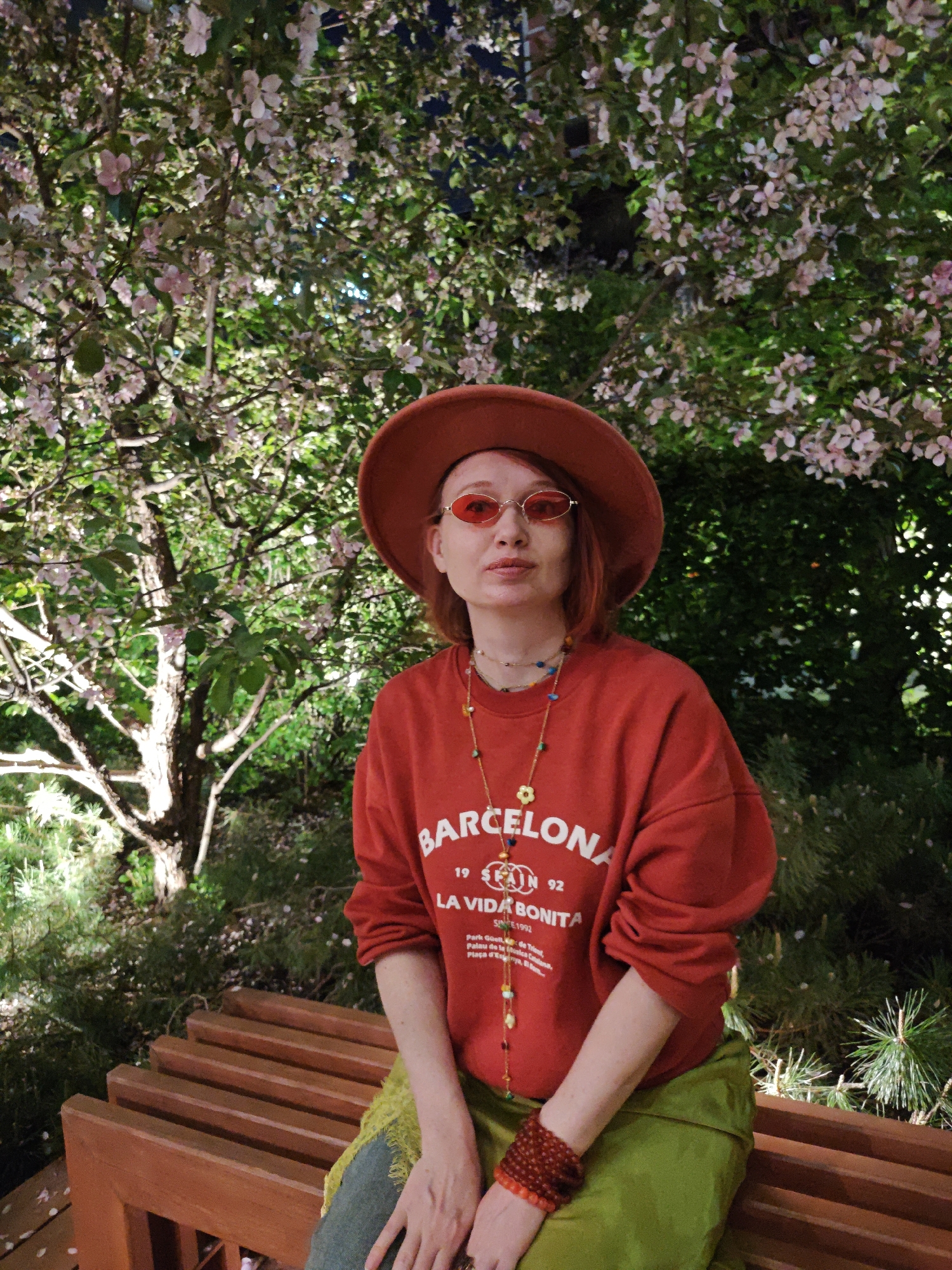 A woman is sitting on a bench under a flowering tree. She is wearing a red hat, red-tinted glasses, a red sweatshirt with "Barcelona" printed on it, a green skirt, and a beaded necklace. She has reddish-brown hair and is looking directly at the camera. The bench is made of wood slats. The tree is covered in small pink flowers. The background appears to be a park-like setting with other trees and plants. The location is not identifiable as Yekaterinburg, Russia, without additional context or visual clues.
