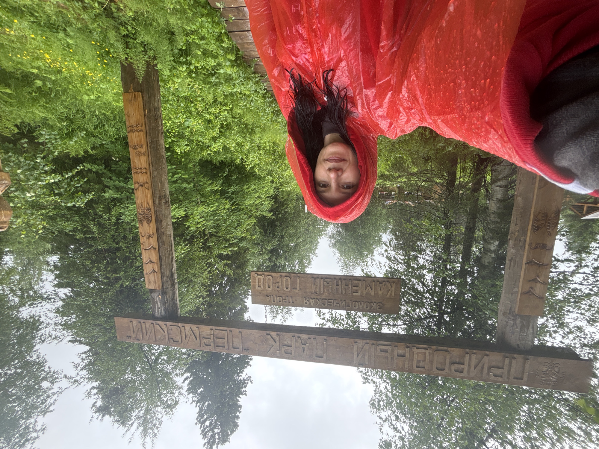 A woman in a red hooded poncho stands in front of a wooden archway sign. The sign, carved in Russian, reads "ПРИРОДНЫЙ ПАРК "ПЕРМСКИЙ"" on the top plank and "ЭКОЛОГИЧЕСКАЯ ТРОПА КАМЕННЫЙ ГОРОД" on the lower plank. The archway is situated in a lush green forest with trees and dense foliage. The ground appears to be a wooden boardwalk. The sky is overcast, suggesting a cloudy or rainy day, and water droplets are visible on the woman's poncho. The location is the Permsky Nature Park, specifically the ecotrail to "Kamenniy Gorod" (Stone City) near the settlement of Usva, Russia.