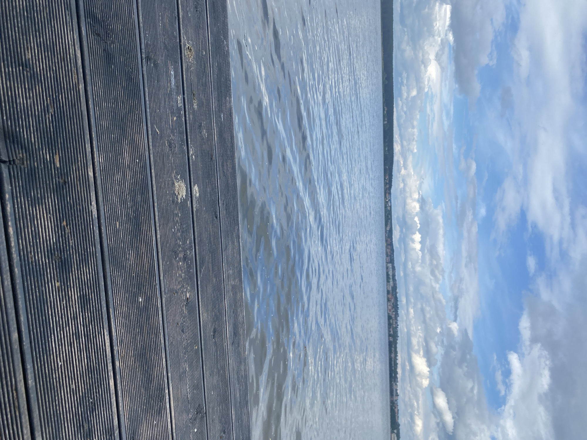 A view of the Votkinskoye Reservoir from a wooden pier. The pier, made of dark, ribbed planks, occupies the foreground and lower portion of the image. Beyond the pier is a vast expanse of calm water, reflecting the sky above. In the distance, a tree-lined shore is visible, with a town or city built along its edge. The sky is partly cloudy, with patches of blue sky interspersed with white and grey clouds. The overall scene depicts a tranquil waterscape.