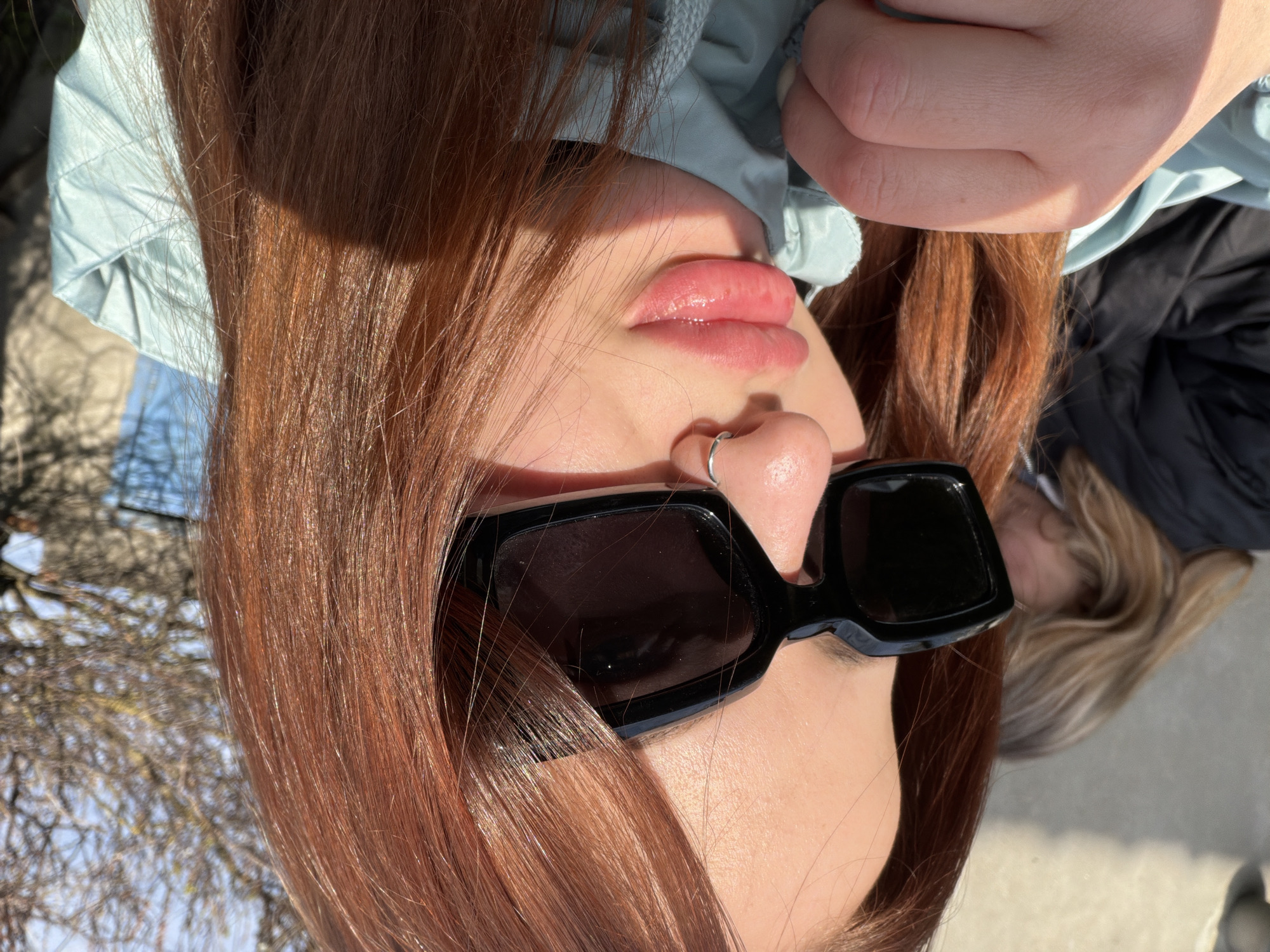 A tilted, close-up portrait shows a person with red hair, wearing black sunglasses and a silver nose ring. Their lips are visible, appearing glossy. Part of a light blue jacket is visible at the top of the frame. The background shows a blurred outdoor environment, including bare tree branches and possibly another person with blonde hair, suggesting an urban setting. The image does not provide sufficient detail to confirm a specific location like Novopokrovskaya, Russia.
