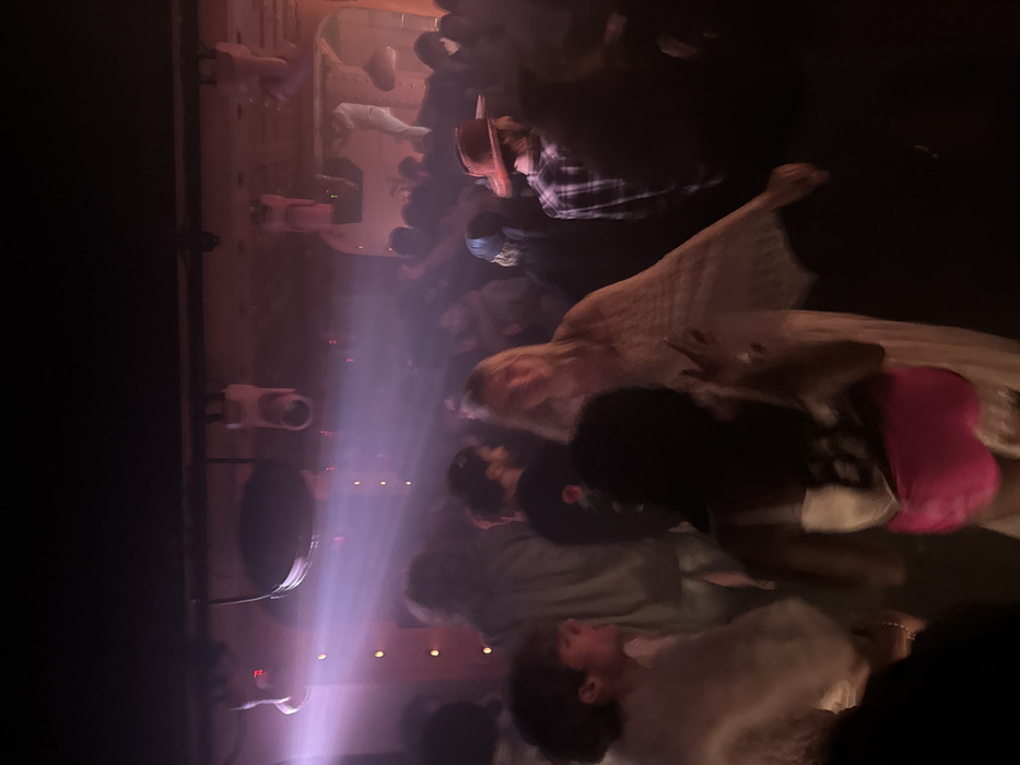 A dimly lit indoor gathering shows numerous individuals. Some wear hats, while one attendee sports a pink garment. The scene is illuminated by bright light beams and interspersed with small decorative lights. The lighting casts shadows and creates a hazy ambiance throughout the room. Cameras are visible along a wall.