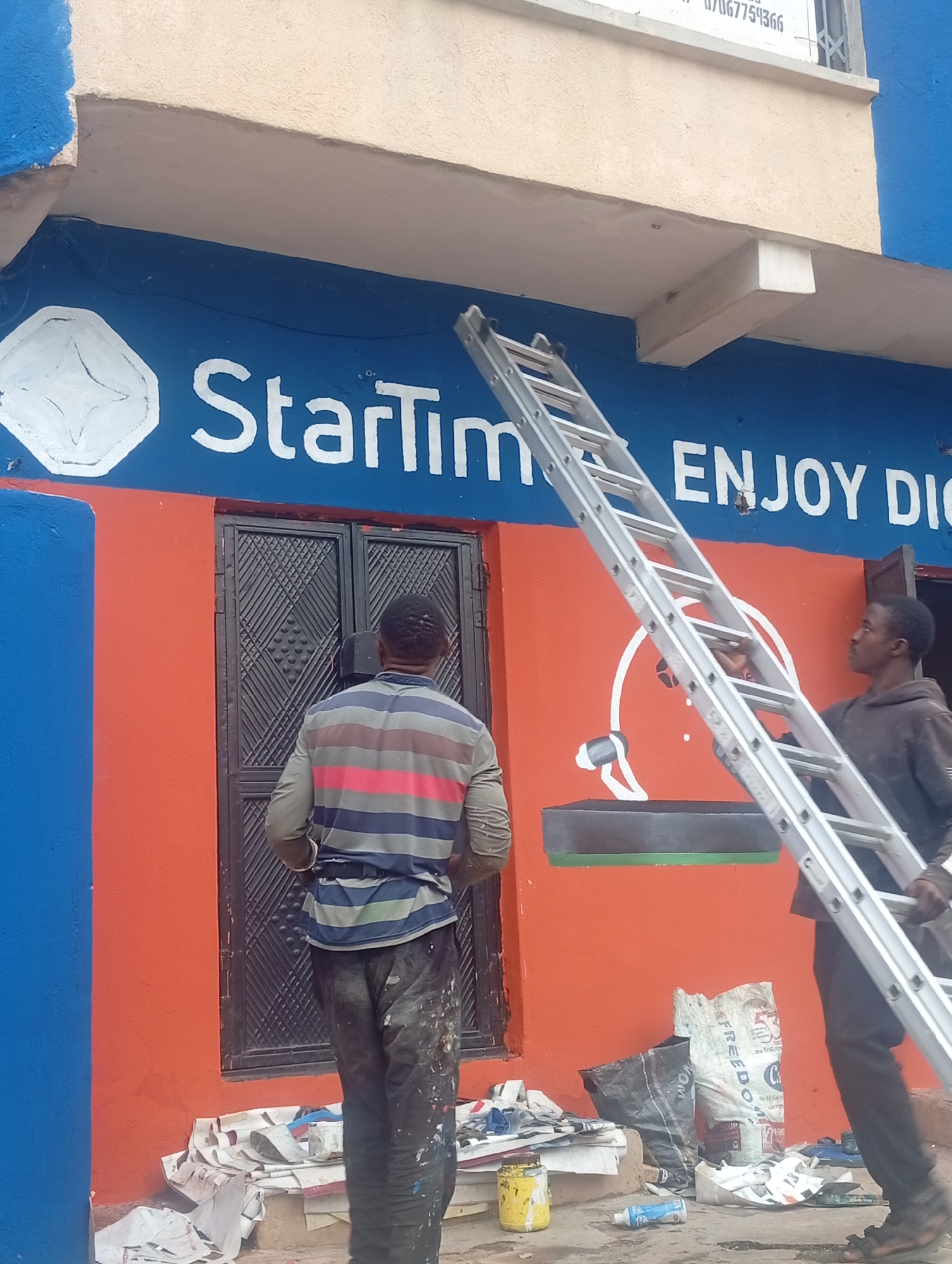 The image captures an outdoor scene likely in Jos, Nigeria, where two men are engaged in painting a building's facade. The building features a vibrant orange lower section and a blue upper section. Prominently displayed on the blue section is the "StarTimes" logo, a white geometric design resembling a star within a diamond shape, followed by white lettering that reads "ENJOY DIGI".
One man, seen from the back, stands in front of a dark-colored double door on the orange section. He wears a horizontally striped shirt with blue, gray, and pink bands, and dark, paint-splattered pants. He appears to be overseeing the painting work.
The second man is positioned on the right side of the image, holding a large, silver, extendable ladder. He wears a dark hooded sweatshirt and dark pants, also with paint splatters. He is looking towards the upper part of the building, likely preparing to climb the ladder or assist with the painting at a higher level.
The ground in front of the building is littered with various materials related to painting, including discarded newspapers, white plastic sheeting, a yellow paint can, a spray can, and other debris. The overall impression is that of an active construction or renovation site, possibly a commercial establishment, given the prominent branding.