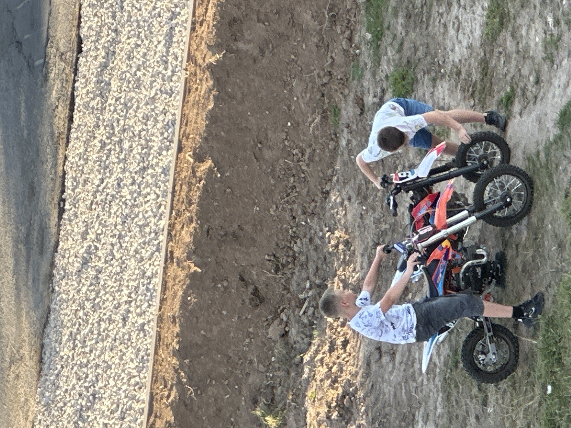 Two young boys are interacting with dirt bikes in a dirt lot in Podolsk, Russia. The boy on the left, wearing a white t-shirt with a blue pattern and dark shorts, is standing next to a dirt bike. The boy on the right, wearing a white t-shirt and blue shorts, is bent over, appearing to adjust something on his dirt bike. The bikes are brightly colored with red, orange, and blue accents. The ground is composed of dirt and gravel, with some sparse vegetation. In the background, a textured surface of light-colored gravel is visible, separated from the dirt lot by a distinct edge. The overall lighting suggests it is daytime, possibly late afternoon due to the warm tone of the light on the gravel.