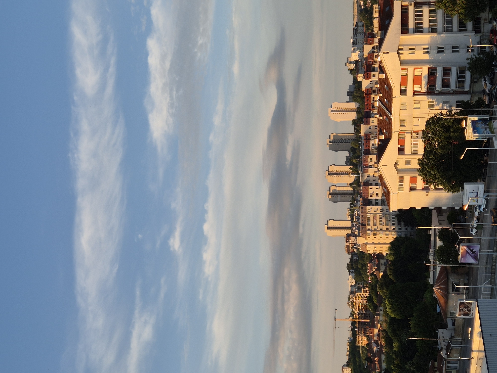The image depicts a cityscape during the golden hour, with the sun casting warm light on buildings and trees. In the foreground, a white building with red accents and numerous windows dominates the lower right portion of the frame. To its left, smaller buildings with red-tiled roofs are visible, nestled among lush green trees. Further in the background, a cluster of taller, light-colored apartment buildings stands out against the sky. A construction crane is positioned on the left side of the mid-ground, suggesting ongoing development. The sky above is a gradient of blue, with wispy clouds tinged with pink and orange from the setting sun. The scene appears to be an urban residential area, likely in the evening.