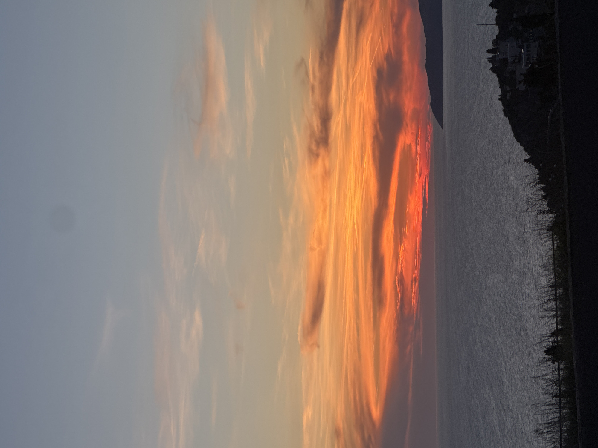 The image depicts a sunset over the Adriatic Sea in Golubovići, Montenegro. The sky is a gradient of colors, transitioning from light blue to orange and red near the horizon. Clouds are visible, illuminated by the setting sun. The sea is a dark gray color. A shoreline with vegetation and a few buildings is visible on the right side of the image. A road runs along the right edge of the scene.