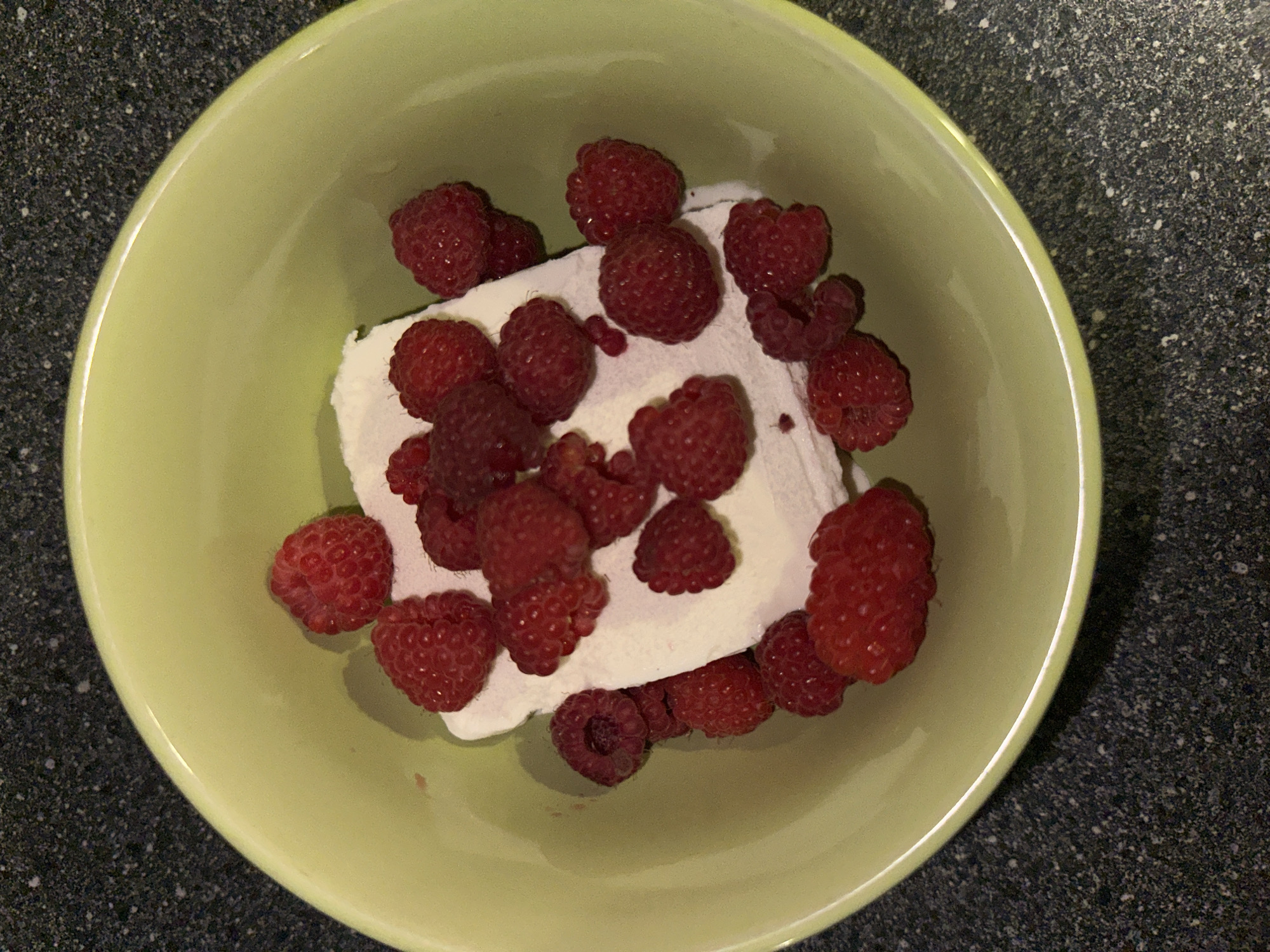 A close-up, top-down shot shows a light green bowl filled with a white substance topped with raspberries. The bowl rests on a speckled, dark gray surface. The raspberries are scattered across the white topping, with some also nestled around the edges of the bowl. The white substance appears to be a creamy dessert, possibly ice cream or whipped cream. The raspberries are a vibrant red color, with visible drupelets. The lighting is somewhat dim, creating subtle shadows. There are no people or any indication of a specific location within the city of Chaikovsky, Russia, or any city.