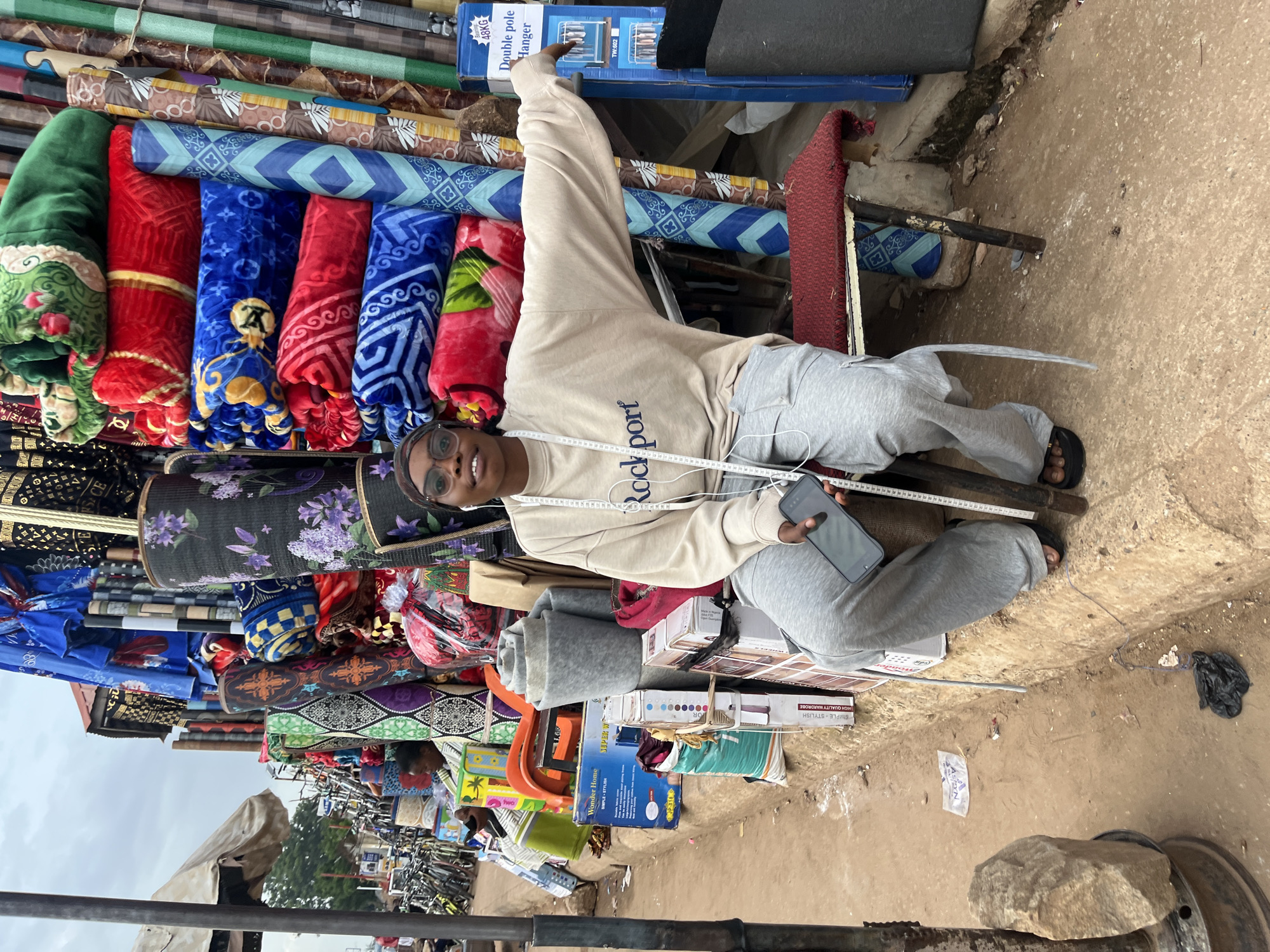 In the city of Bukuru, Nigeria, a young woman wearing a light-colored sweatshirt and grey sweatpants sits on a red-cushioned stool in front of a stall overflowing with textiles. The stall displays a variety of rolled fabrics, including blankets and patterned materials in vibrant colors such as red, blue, green, and purple, as well as various types of baskets and household items like a blue box labeled "Wonder Home." The woman is holding a smartphone and has a measuring tape draped around her neck. She appears to be interacting with the camera, smiling slightly and looking towards it. The environment suggests a bustling market scene, with other stalls and bicycles visible in the background. The ground is made of dirt and concrete, with some scattered debris.