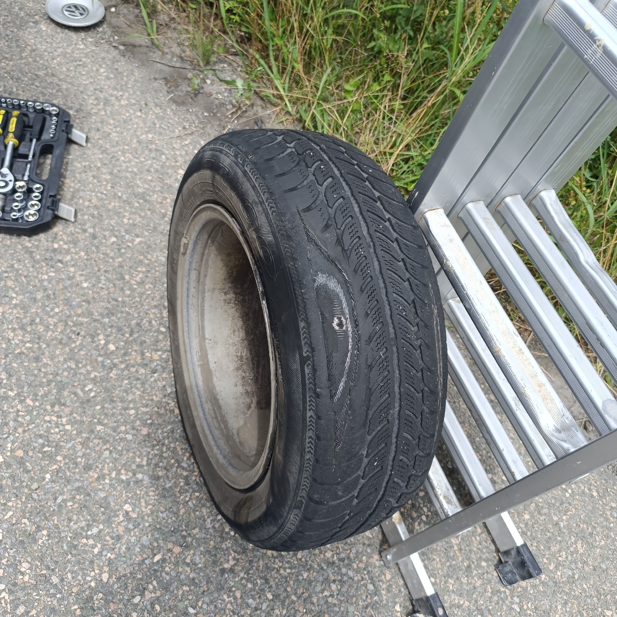 A flat tire is shown on a paved surface next to a silver ladder. A set of tools is to the left of the tire, and a silver Volkswagen hubcap is in the upper left corner. Green grass is visible in the background.