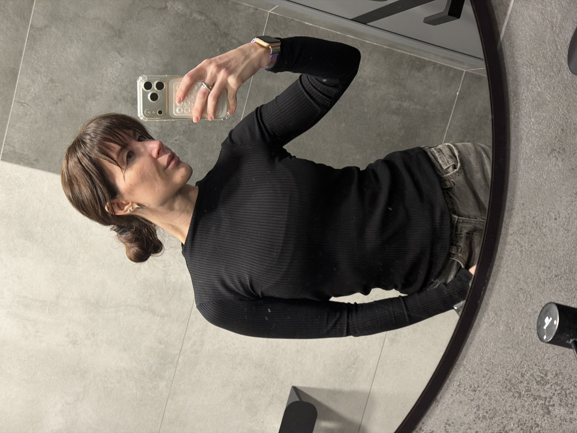A woman with brown hair and bangs, tied back in a bun, is captured taking a mirror selfie in Perm, Russia. She is reflected in a dark-framed, oval mirror, positioned in what appears to be a bathroom or dressing area. She wears a black, ribbed long-sleeved top and gray or dark-wash denim jeans.
The woman is holding a smartphone, which appears to be an iPhone with a clear case showcasing a triple camera lens setup, pointed towards her reflection to capture the image. On her left hand, she wears a silver ring on her ring finger, and a smart watch with a colorful, possibly braided, band is visible on her left wrist. Small silver hoop earrings adorn her ears. She looks intently at the phone screen, maintaining a neutral expression.
The background visible in the mirror's reflection consists of gray tiled walls and floor, with subtle grout lines. Above her shoulder, a dark fixture that might be part of a shower or a towel rack is partially visible. To the right of the mirror, a dark, cylindrical object with some indistinct white markings is present on the wall. The lighting appears to be indoor and even. No clear, readable text is visible on any objects in the image.