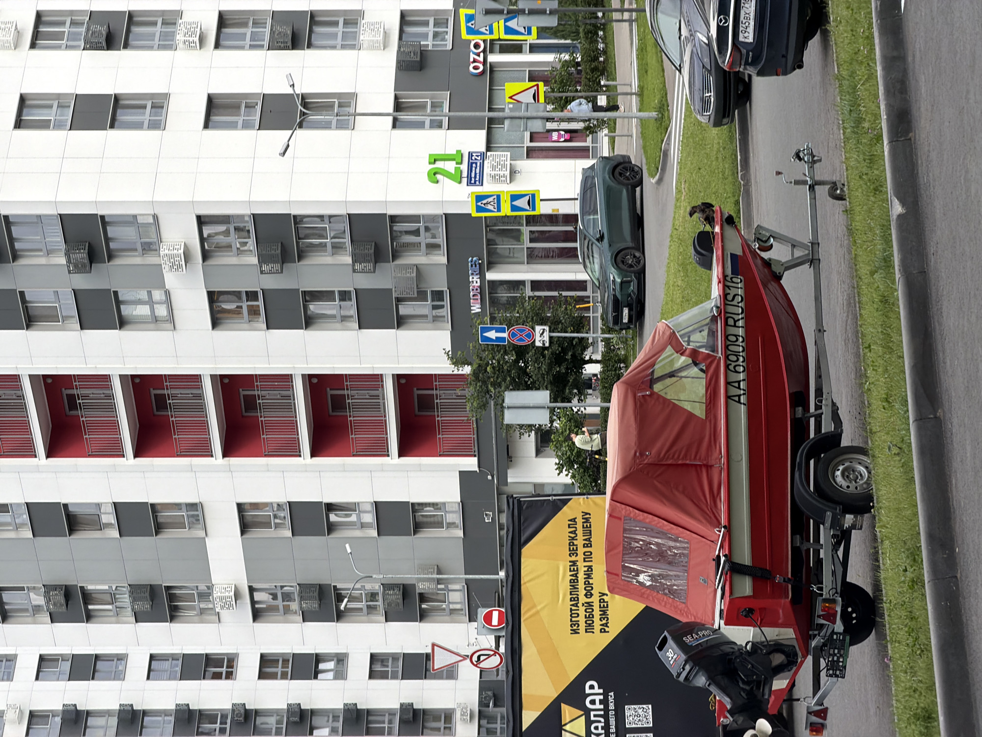 The image captures a scene in Kazan, Russia, featuring a residential building and a boat on a trailer parked on the side of a road. The building is modern with a white facade and balconies with red accents, punctuated by numerous windows and gray square panels. Several signs are visible on the building, including "OZO" and "WILDBERRIES," suggesting commercial establishments on the ground floor. Traffic signs, including a yield sign and a no-entry sign, are also present.
The foreground is dominated by a red boat mounted on a trailer. The boat has a canopy and an outboard motor labeled "SEA-PRO." The license plate on the trailer reads "AA 6909 RUS 16." A bird, possibly a hawk or eagle, is perched on the boat. To the right of the boat and trailer, a dark-colored car is parked on the asphalt road, with another dark car further down the street. A lush green grassy area borders the road.
The overall scene appears to be set during the daytime, under natural light. The weather seems pleasant, as indicated by the greenery and the outdoor setting. The presence of cars, a boat, and a building suggests an urban or suburban environment. No specific event is clearly depicted, but the setup implies a recreational or transport-related activity. The text visible on the building and the boat provides brand names and a license plate number, along with a slogan in Russian on a large banner, "ИЗГОТАВЛИВАЕМ ЗЕРКАЛА ЛЮБОЙ ФОРМЫ ПО ВАШЕМУ РАЗМЕРУ" which translates to "WE MANUFACTURE MIRRORS OF ANY SHAPE ACCORDING TO YOUR SIZE."